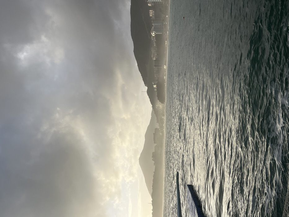 The image captures a serene coastal landscape during what appears to be a late afternoon or early morning, with soft, diffused sunlight. The photograph is oriented vertically, but when viewed horizontally, it depicts a wide expanse of ocean on the right, meeting a mountainous coastline on the left.
The sky, occupying the upper left portion of the image, is mostly overcast with a thick layer of grey and white clouds. There are hints of brighter light breaking through the clouds, suggesting the sun is either rising or setting, or obscured but strong. This creates a soft, almost ethereal glow in the atmosphere, particularly near the horizon line where the sky meets the mountains.
The landmass on the left consists of several layers of mountains or hills, receding into the distance. The closest mountain is dark and silhouetted against the bright sky, showing some irregular peaks and valleys. On the slopes of these mountains, especially the closer ones, several buildings are visible. These appear to be multi-story structures, possibly apartments or hotels, nestled along the coastline. Their lights are either on or reflecting the ambient light, appearing as tiny specks of brightness. A faint haze or mist seems to cling to the lower slopes of the mountains and the coastline, further softening the distant details. A tall, thin structure, possibly a communication tower or flagpole, can be seen on one of the mountain ridges.
The body of water, likely an ocean or a large bay, dominates the right side of the image. Its surface is rippled with numerous small waves, creating a textured pattern across its expanse. The water reflects the soft, bright light from the sky, resulting in streaks and patches of shimmering silver and grey. The light glints off the wave crests, highlighting their movement. In the bottom right corner, a dark, partially submerged structure extends into the water. It appears to be a pier, jetty, or a series of wooden or metal planks, with visible gaps between the individual components. The structure is mostly in shadow but its upper surface catches some light. There are no visible people or discernible text in the image.