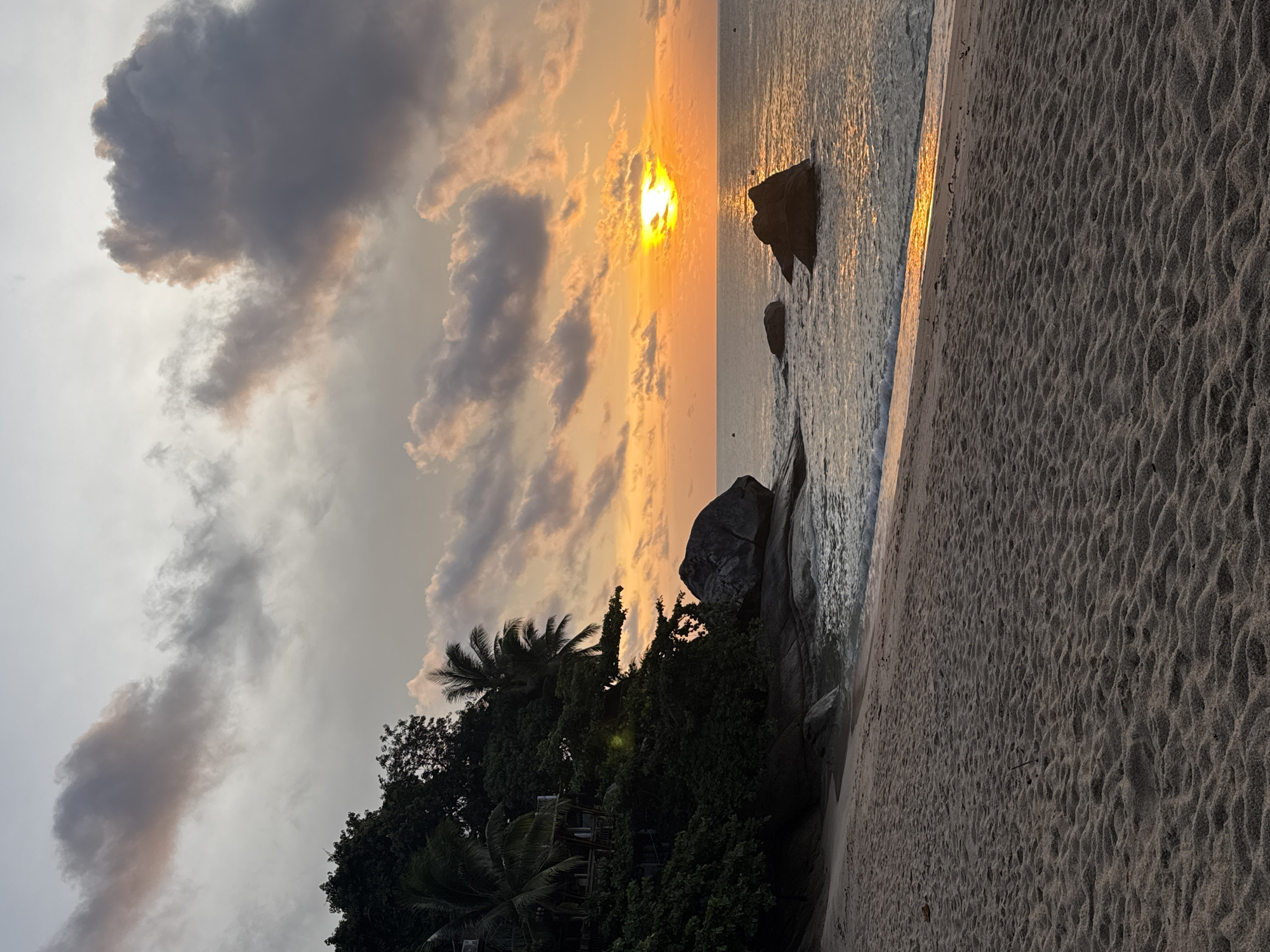 The image depicts a beach at sunset in Baan Thong Nang, Thailand. Dark, heavy clouds occupy the upper left of the frame, contrasting with the vibrant orange and yellow hues of the setting sun. The sun is partially obscured by clouds, casting a golden reflection on the water. Several large rocks are visible in the sea near the shoreline. Lush green vegetation, including palm trees, lines the beach in the lower-left portion of the image. The foreground is dominated by sandy beach with footprints and ripple patterns.