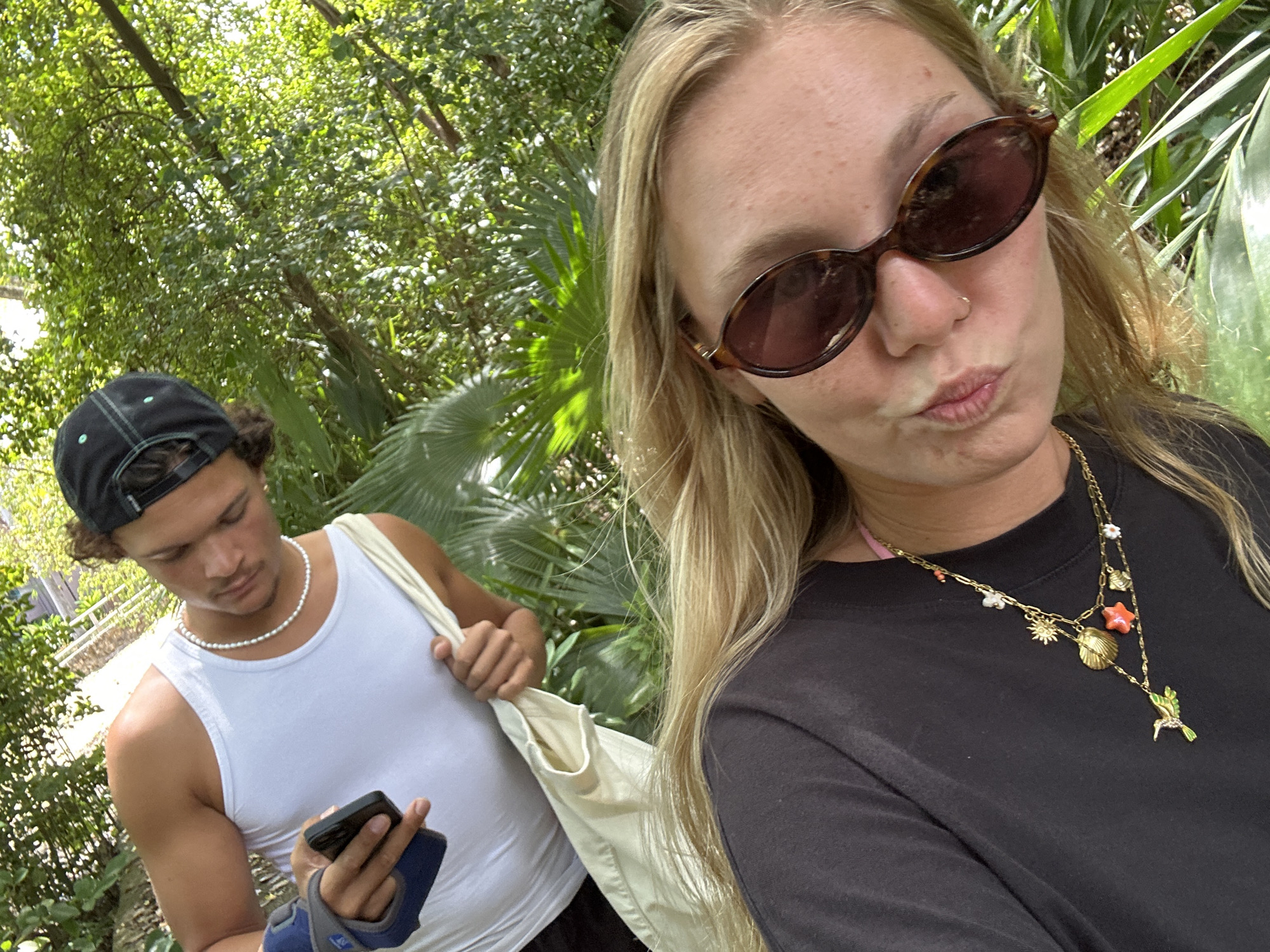 Two people are captured in this medium shot. A woman is in the foreground, wearing brown sunglasses, a black t-shirt, and a multi-strand gold necklace with various charms. Her expression is a pucker. To her left and slightly behind, a man wears a white tank top, a pearl necklace, and a black baseball cap. He's looking down at a mobile phone held in his left hand. He has a white tote bag over his left shoulder. The background is filled with green foliage, suggesting a park or garden setting. Based on the location, the scene is most likely in Sainte-Anne, France.