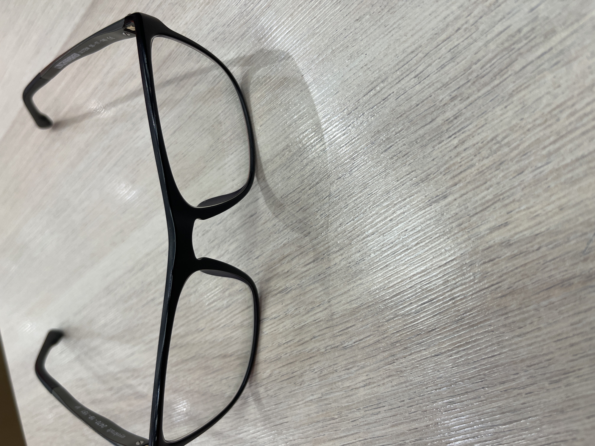 A pair of black-rimmed eyeglasses with clear lenses is placed on a light-colored wooden surface with a visible wood grain pattern. The eyeglasses are positioned slightly angled, with the temples extended outward. The left temple has some text engraved on it, which appears to be "eyekraft 2470-19-151 M". The right temple also has engraved text, visible as "ULTEM 55-17-140 CE". The surface beneath the eyeglasses shows natural light reflection, creating a subtle shadow. The background is out of focus, suggesting a shallow depth of field. The image is taken from a top-down perspective. There are no people or animals present. The location is not specified as Moscow, Russia.