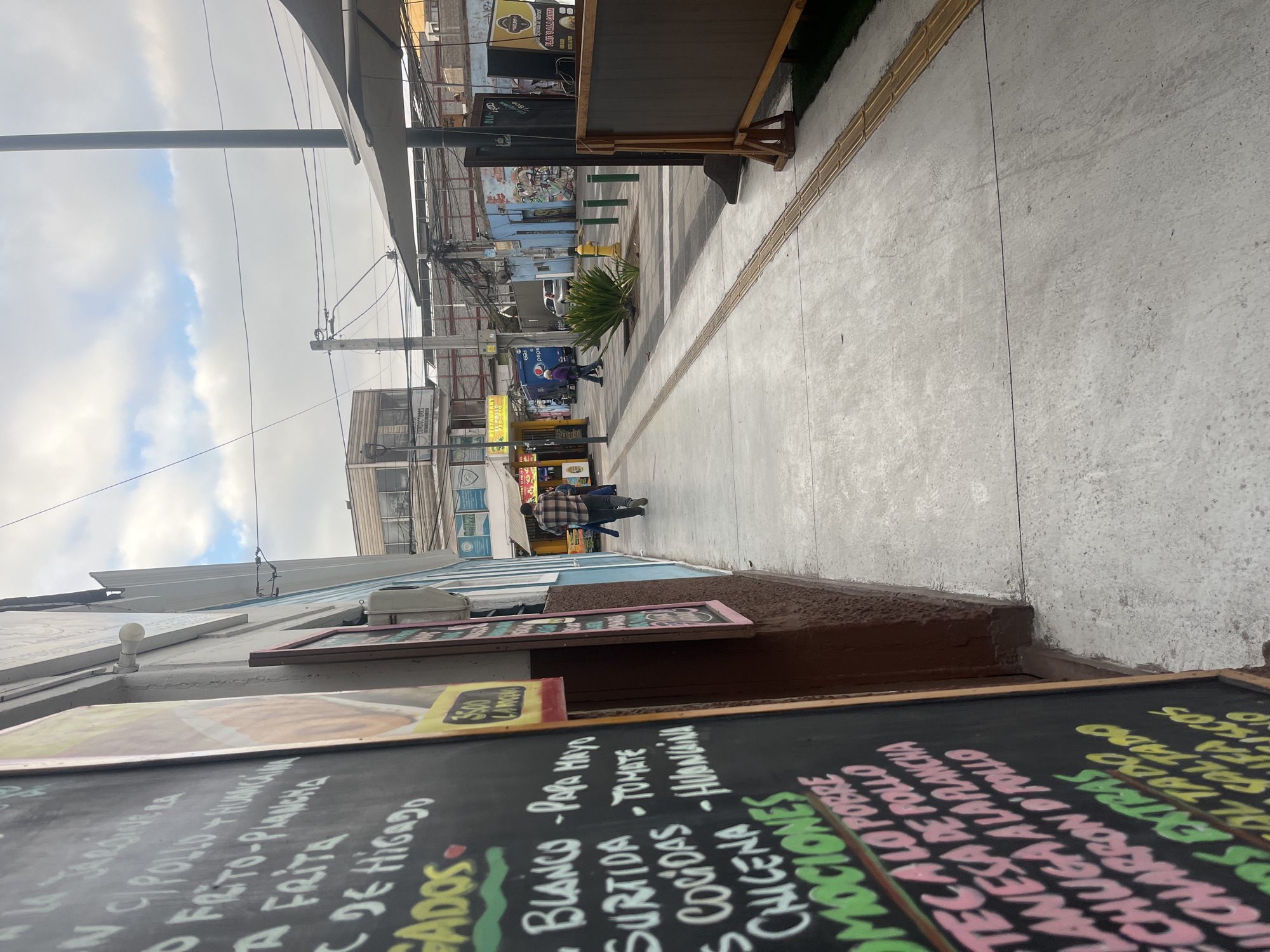 The image shows a sidewalk with a blackboard menu in the foreground, on the left side of the frame. The blackboard lists food items and prices in Spanish. In the background, there is a street with several buildings, shops, and a few people walking. A Pepsi truck is visible on the street, as well as a fire hydrant and some streetlights. The sky is cloudy. The location is identified as Iquique, Chile, based on the visible text on the blackboard and surrounding context. The scene appears to be a commercial area with outdoor dining or food stalls.