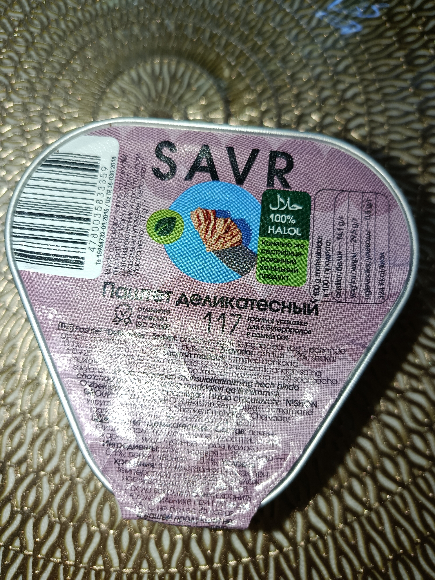 The image shows a close-up of a food product container, likely a can or a small tub. The container is predominantly purple and silver, with text and graphics printed on its surface.
Prominently featured is the brand name "SAVR" in large, dark letters. Below this, there's a green certification logo with Arabic text that translates to "Halal" and "100% HALAL" written underneath in English.
To the right of the brand name, there is a nutritional information section presenting data per 100g of product. This includes:
* oqsillar/belki (proteins/fats): 14.1 g/r
* yog'lar/жиры (oils/fats): 29.5 g/r
* uglevodlar/углеводы (carbohydrates): 0.5 g/r
* 324 Kkal/Kкал (kilocalories)
On the left side of the container, there's a barcode with the number "4780003583359" and additional text including "Ts 15964732-01:2015 / Uz TR 36-030:2018".
There is also text in Russian, stating "Паштет деликатесный" which translates to "Delicatessen pate". Below this, the number "117" is visible.
Further text, appearing to be a list of ingredients and other product information, is present in a smaller font in various languages, including Russian and possibly Uzbek. This section includes details about the product's composition, such as the presence of "0,1% пепсин" (0.1% pepsin) and "0,1% слизи" (0.1% mucus). It also mentions storage conditions, like "tребует ог 0°C to +25°C" (requires from 0°C to +25°C) and "не более 48 soot`gacha" (no more than 48 hours). The text "48 soot'gacha" might refer to a duration in hours or a unit of time. There is also mention of "Massa Neto: 117 g/r" (Net Mass: 117 g/r).
The environment appears to be indoors, with the container placed on a textured, reflective surface that creates a patterned background of light and shadow. The surface's texture resembles a woven or patterned fabric, possibly a tablecloth or a decorative mat. The lighting suggests a warm, artificial light source.
There are no people or any discernable city elements of Tashkent, Uzbekistan, visible in the image. The context provided by the text ("Uz TR 36-030:2018" likely refers to Uzbekistan's Technical Regulations) suggests the product is either manufactured in or intended for the Uzbek market.