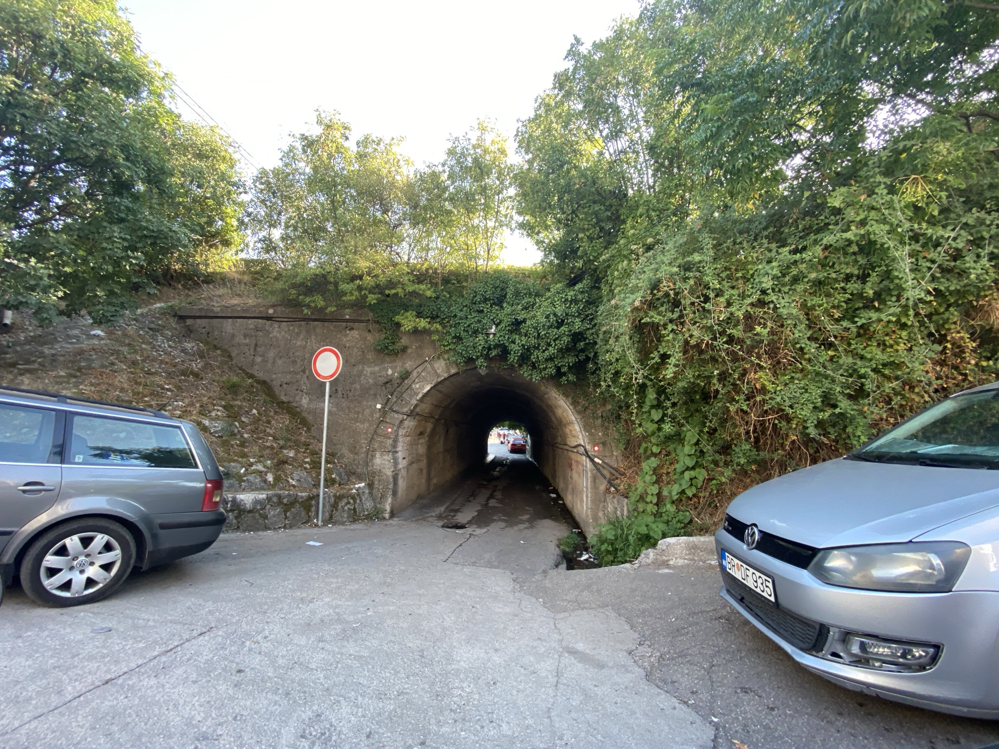 In Zankovići, Montenegro, a two-lane road leads to a stone tunnel entrance. A gray Volkswagen wagon is parked on the left side of the road, facing away from the tunnel. A silver Volkswagen Polo is parked on the right side, facing the tunnel entrance. The tunnel is illuminated by two red lights on the left and right walls, and natural light from the other end. Vegetation covers the walls surrounding the tunnel. A "no entry" sign is posted to the left of the tunnel entrance. The ground is asphalt and concrete, with some debris scattered around.