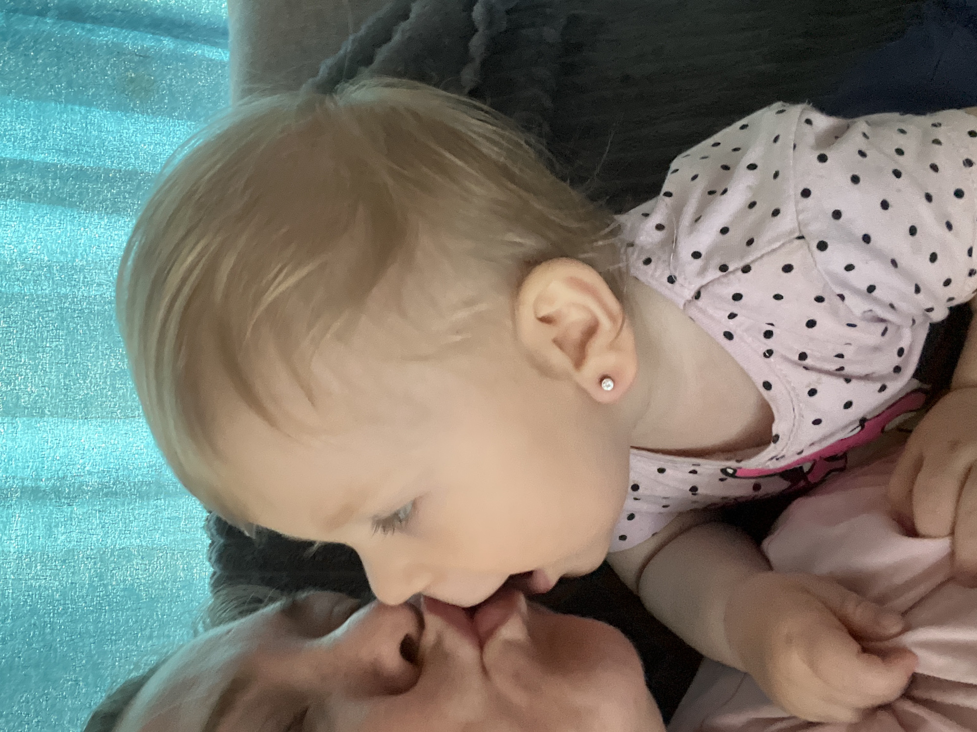 A close-up, outdoor, eye-level, medium shot shows two people, an adult and a baby, in what appears to be a home environment. The adult, on the left, is kissing the baby, who is on the right. The baby has fair skin and blond hair and is wearing a light pink polka-dot shirt. The adult also has fair skin and blond hair. The background is a turquoise textured curtain and a dark grey blanket. The lighting is soft and natural, suggesting it is daytime. The image does not provide any specific information about the location in the city of Fokino, Russia.
