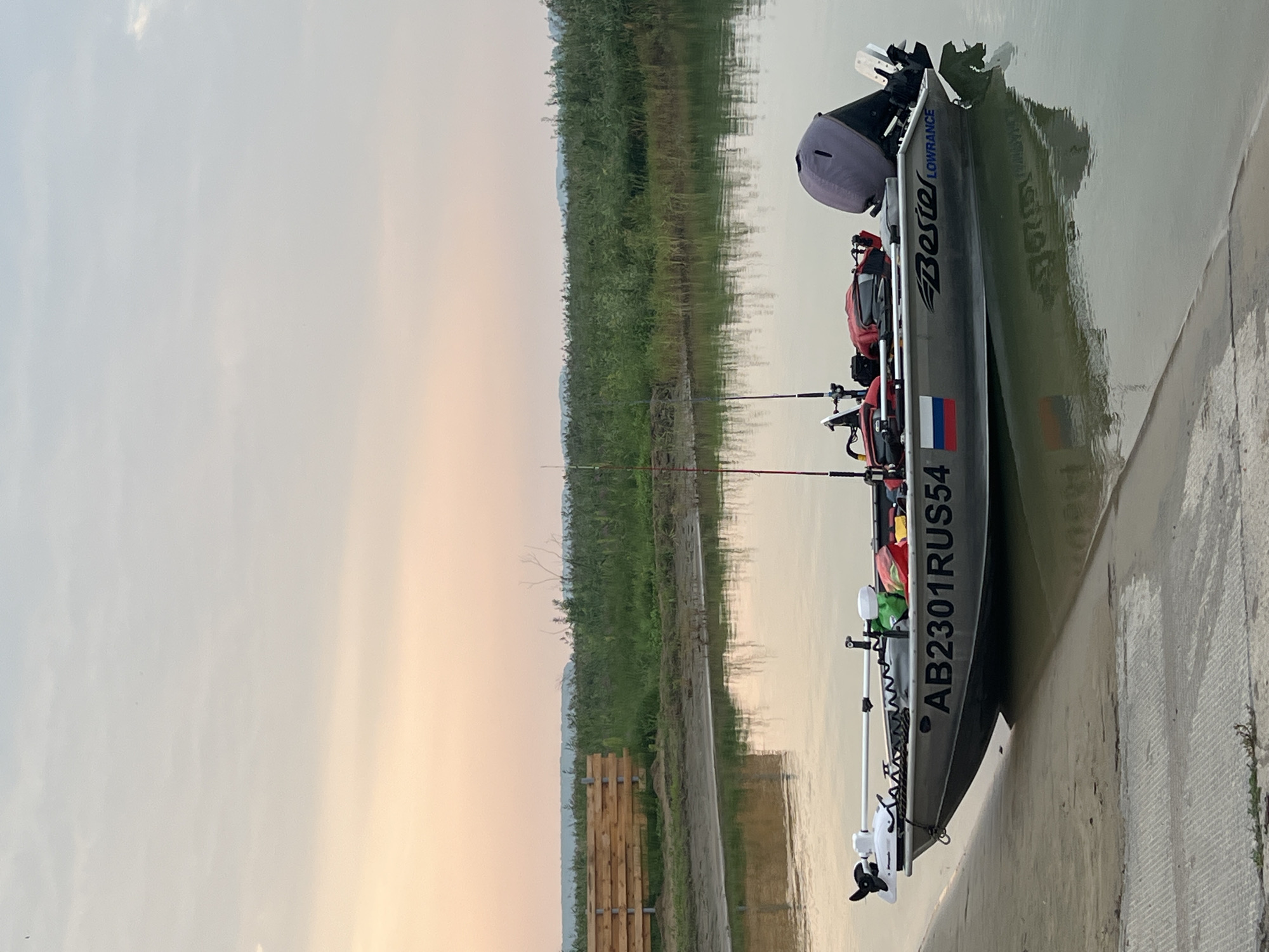 The image shows a silver fishing boat, labeled "AB2301RUS54" and with "Besiter LOWRANCE" branding, docked on a concrete ramp at the edge of a calm body of water. The boat is equipped with two fishing rods, a trolling motor at the bow, and a larger outboard motor at the stern. Several red and green bags are secured within the boat. The background features a grassy embankment with a wooden fence and dense green vegetation stretching towards a hazy horizon. The sky is a soft gradient of pale orange and blue, indicating either sunrise or sunset. The scene appears to be in a rural or natural setting, consistent with the location of Zav'yalovo, Russia, which is known for its lake and natural landscapes. No people are visible in the image.