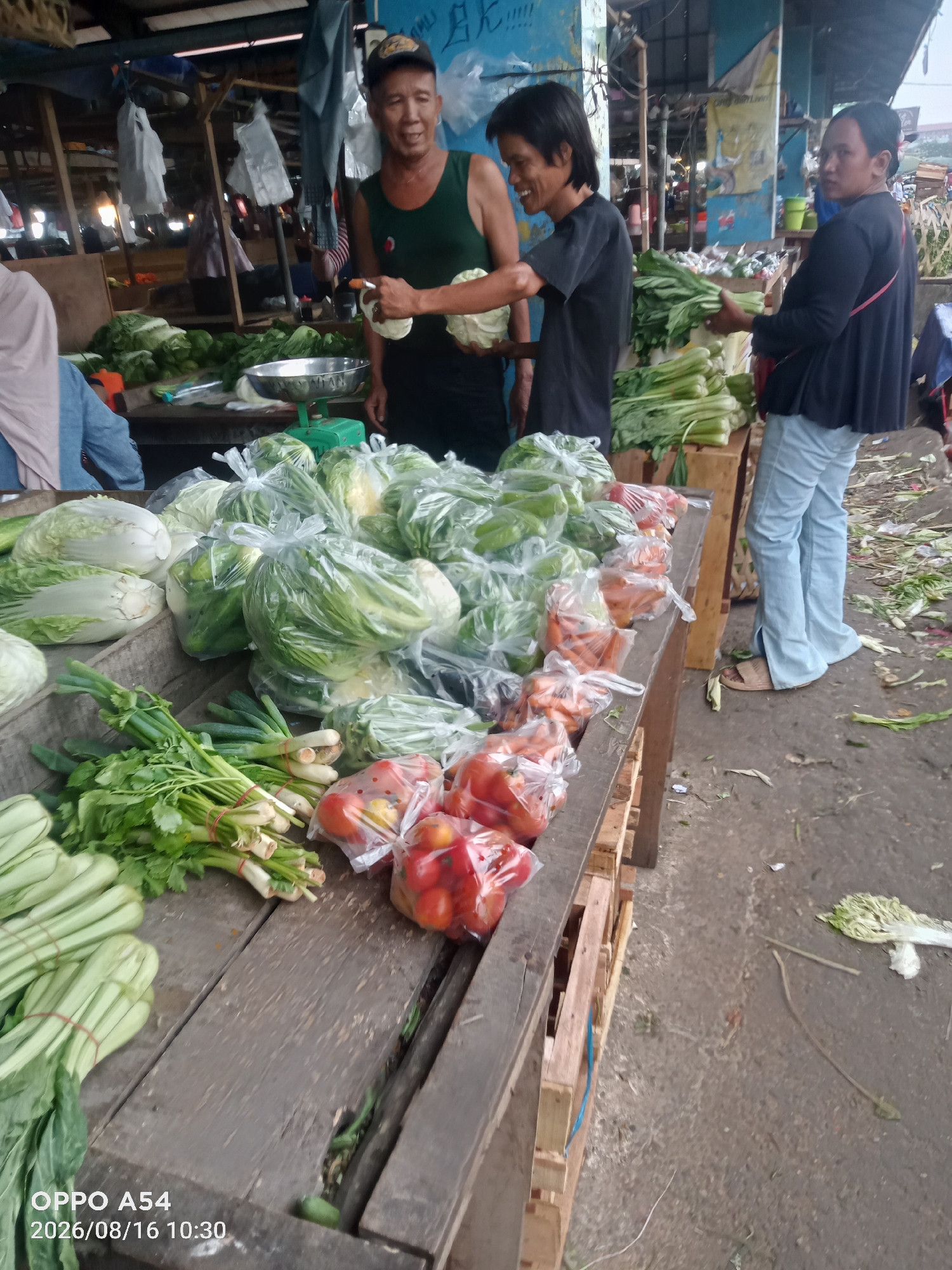 The image depicts a street market scene, likely in Jambi, Indonesia, with an emphasis on fresh produce. Several individuals are present, primarily behind a wooden stall laden with vegetables. In the foreground, a variety of leafy greens, including what appear to be bok choy and scallions, are neatly bundled and arranged. Bags of cabbage and other vegetables are also visible. To the right, a woman in light blue pants and a dark top stands facing away from the camera. Two men are engaged in conversation near the stall. One man, wearing a green tank top and a baseball cap, holds a bundle of vegetables and appears to be showing them to the other man, who wears a black t-shirt. The market environment is suggested by the wooden stalls, tarps, and the rough ground. The time stamp on the image indicates it was taken on August 16, 2026, at 10:30.