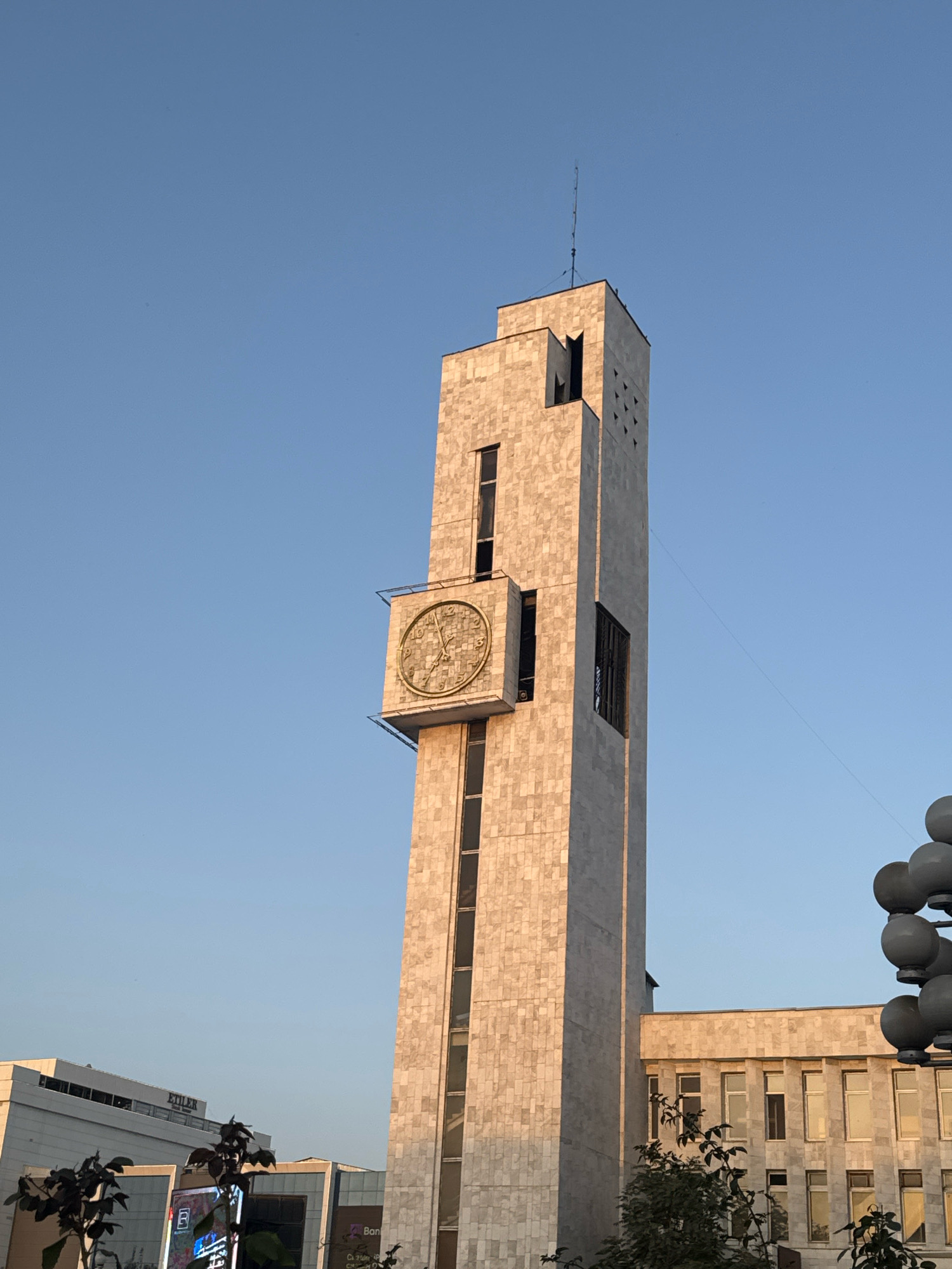 This is a close-up, eye-level, outdoor, daylight, medium shot of the Bishkek City, Kyrgyzstan Clock Tower, situated against a clear blue sky. The tower, constructed of light-colored stone tiles, dominates the frame and is illuminated by warm, late-afternoon sunlight, casting subtle shadows. A prominent circular clock face, adorned with Roman numerals and decorative detailing, is mounted on the tower's left side. Multiple windows of varying shapes and sizes punctuate the tower's facade. To the right of the clock tower, a portion of a lower, longer building, also clad in similar stone tiles, is visible. This building features a row of rectangular windows. In the foreground, the tops of dark green leafy trees and branches partially obscure the view of the lower buildings. The sky is clear and bright, with no clouds present.