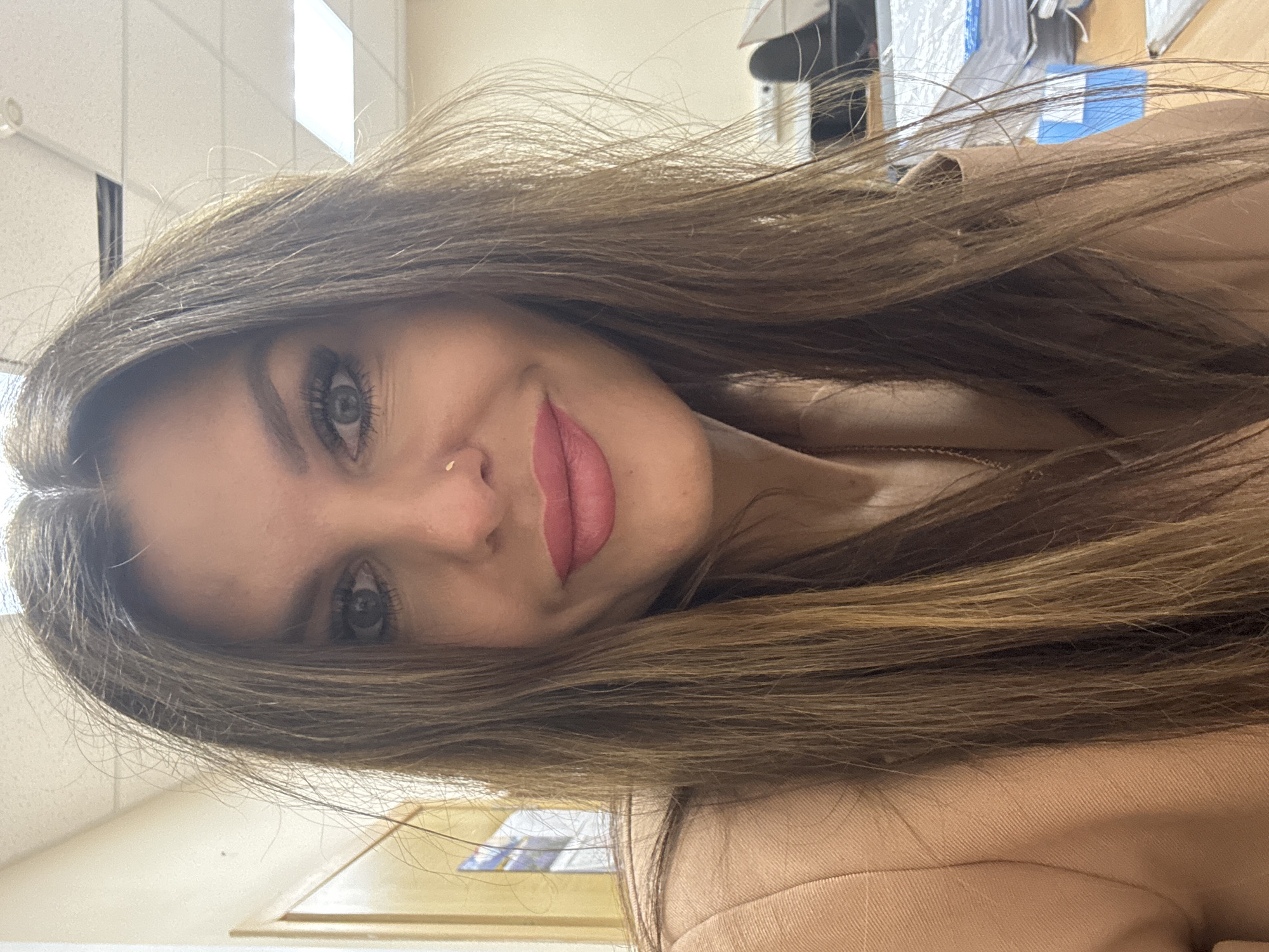 A woman with long, brown hair is facing the camera. She has fair skin, light eyes, and is wearing lipstick. She is wearing a tan jacket or blouse. The background appears to be an office setting with fluorescent lighting. The specific location of Chaikovsky, Russia, cannot be confirmed from the visual data provided.