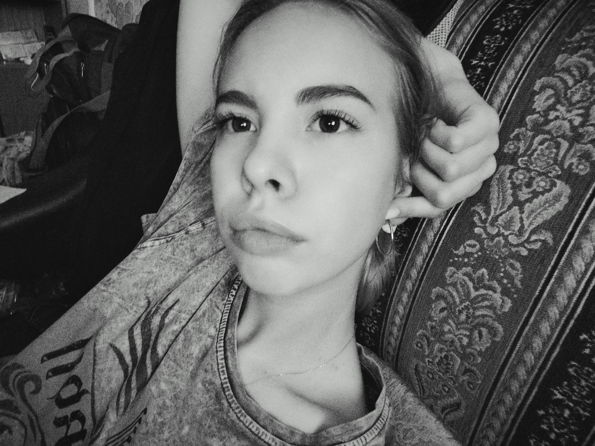 A black and white medium close-up shot shows a young woman with her head tilted to the side and her hand resting on her head. She is looking off to the right of the frame with a neutral expression. She has dark, arched eyebrows and visible eyelashes. She is wearing a t-shirt with a printed design on the sleeve. A thin necklace is visible around her neck. She is leaning against a patterned cushion or sofa. The background is dark and out of focus, with hints of furniture. The lighting appears to be indoor lighting, possibly from a window or lamp. No specific time of day or weather conditions can be determined. The setting is likely a home or indoor space. There is no discernible text in the image.