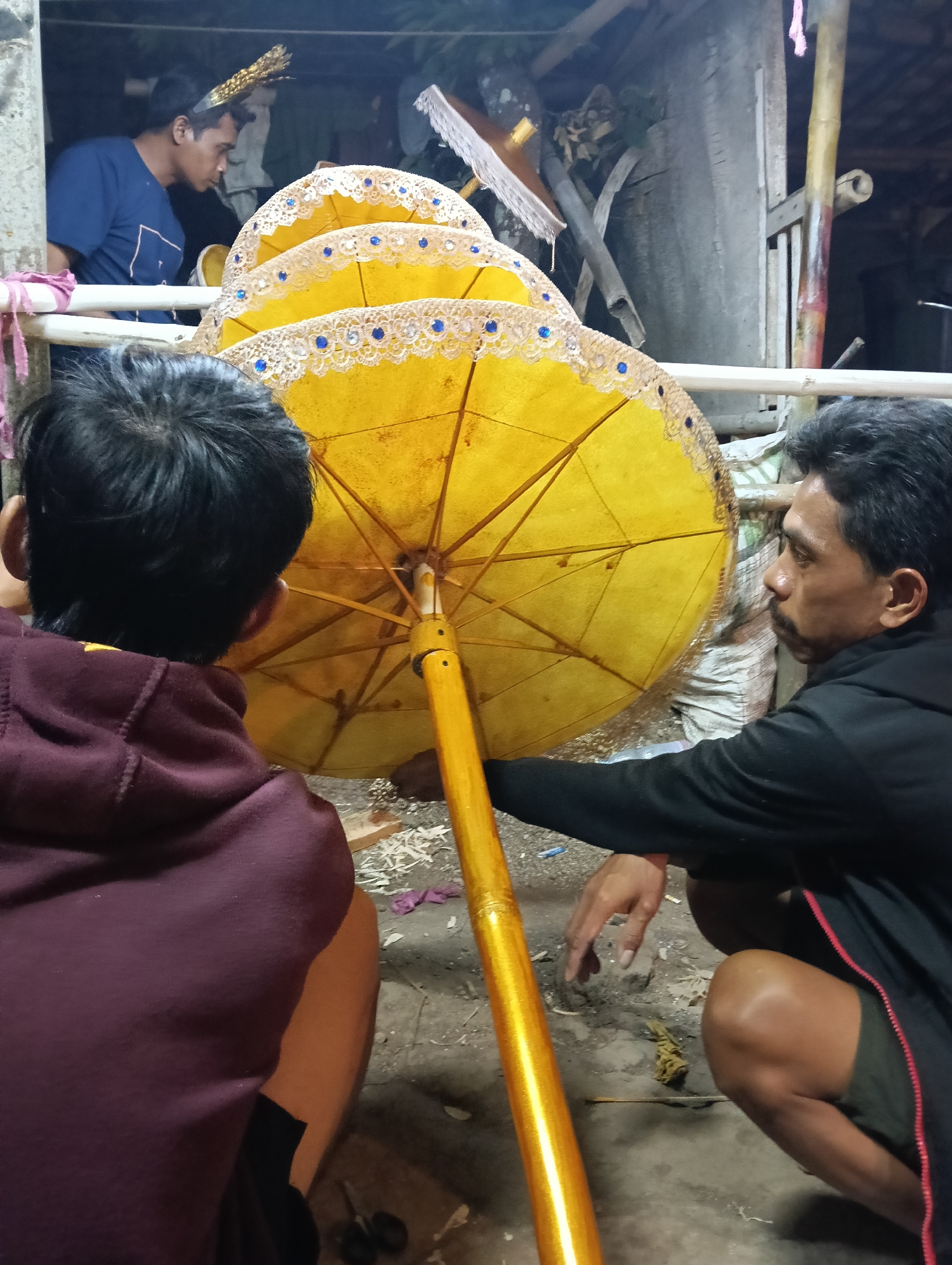 Three individuals are seen in a workshop, likely in Tasikmalaya, Indonesia, as they work on decorative umbrellas. In the foreground, two men are kneeling, their backs mostly to the camera. The man on the right, wearing a black hoodie and green shorts, is actively engaged with a large, bright yellow umbrella, adjusting its golden pole. The umbrella is intricately decorated along its edge with gold trim and small blue beads. To the left, another man, viewed from the back and wearing a maroon hoodie, observes the umbrella. In the background, a third man, wearing a blue t-shirt and a golden headdress, looks on. The environment appears rustic, with unfinished wooden structures, scattered debris, and bamboo poles visible, suggesting a traditional craft setting.