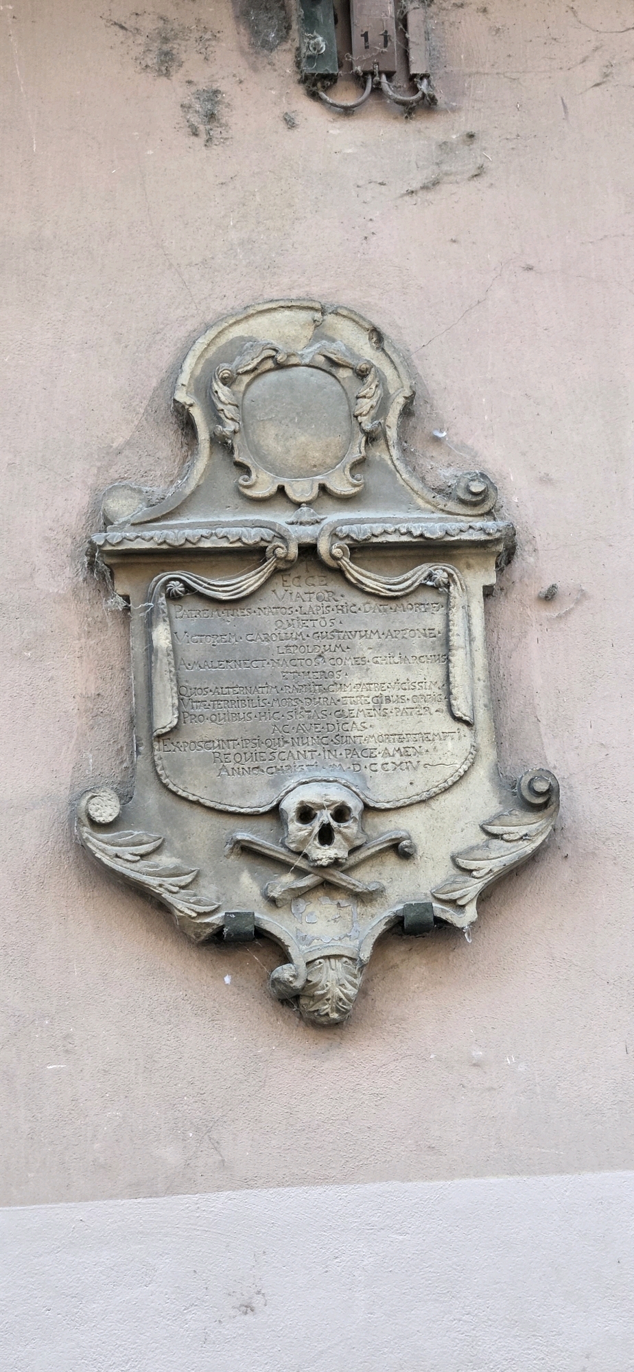 This image shows a sepia-toned stone plaque mounted on a pinkish wall. The plaque is ornate and features a central inscription area surrounded by decorative elements. At the bottom of the plaque, a skull and crossbones are prominently displayed, along with a coat of arms. The inscription is in Latin, with some letters difficult to decipher due to age and weathering. The text appears to be a memorial inscription, possibly for a family or individual, given the presence of names and a date. The wall itself is textured and shows signs of age, with some dirt and cobwebs present, particularly around the plaque and some electrical conduits visible above it. The overall impression is of an old historical marker located in an urban environment.
This plaque is located at 1 rue des Orfèvres, Strasbourg, France.