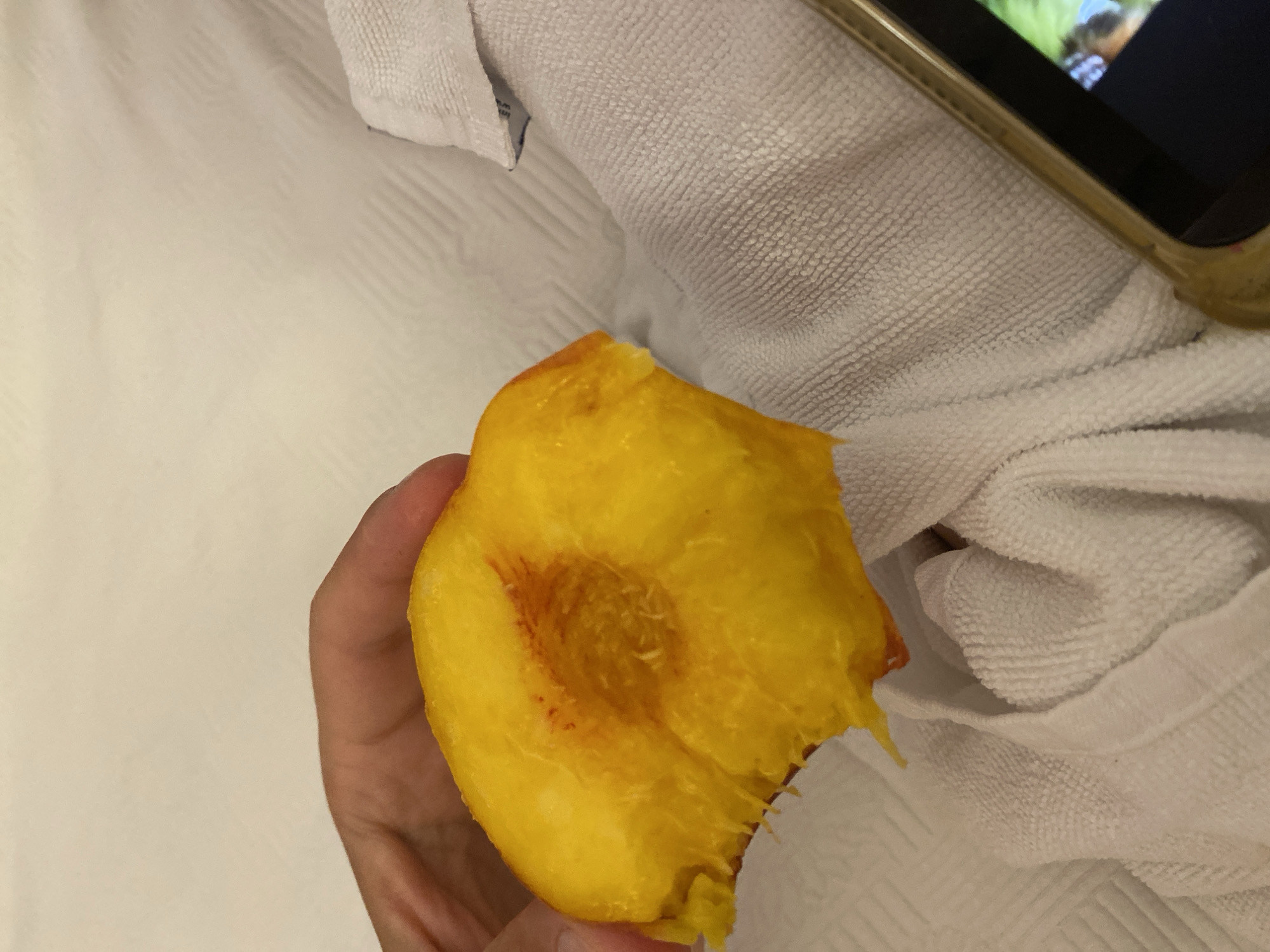A hand holds a partially eaten peach over a white textured surface, likely a towel or bedspread. A portion of a gold-rimmed smartphone with a screen displaying an image is visible in the lower right corner. The scene is illuminated by artificial light, casting soft shadows. The peach is bright yellow with some reddish blush near the core. The background appears to be indoors, with a white, patterned fabric. There is no indication of the city of Aksu, Turkey.