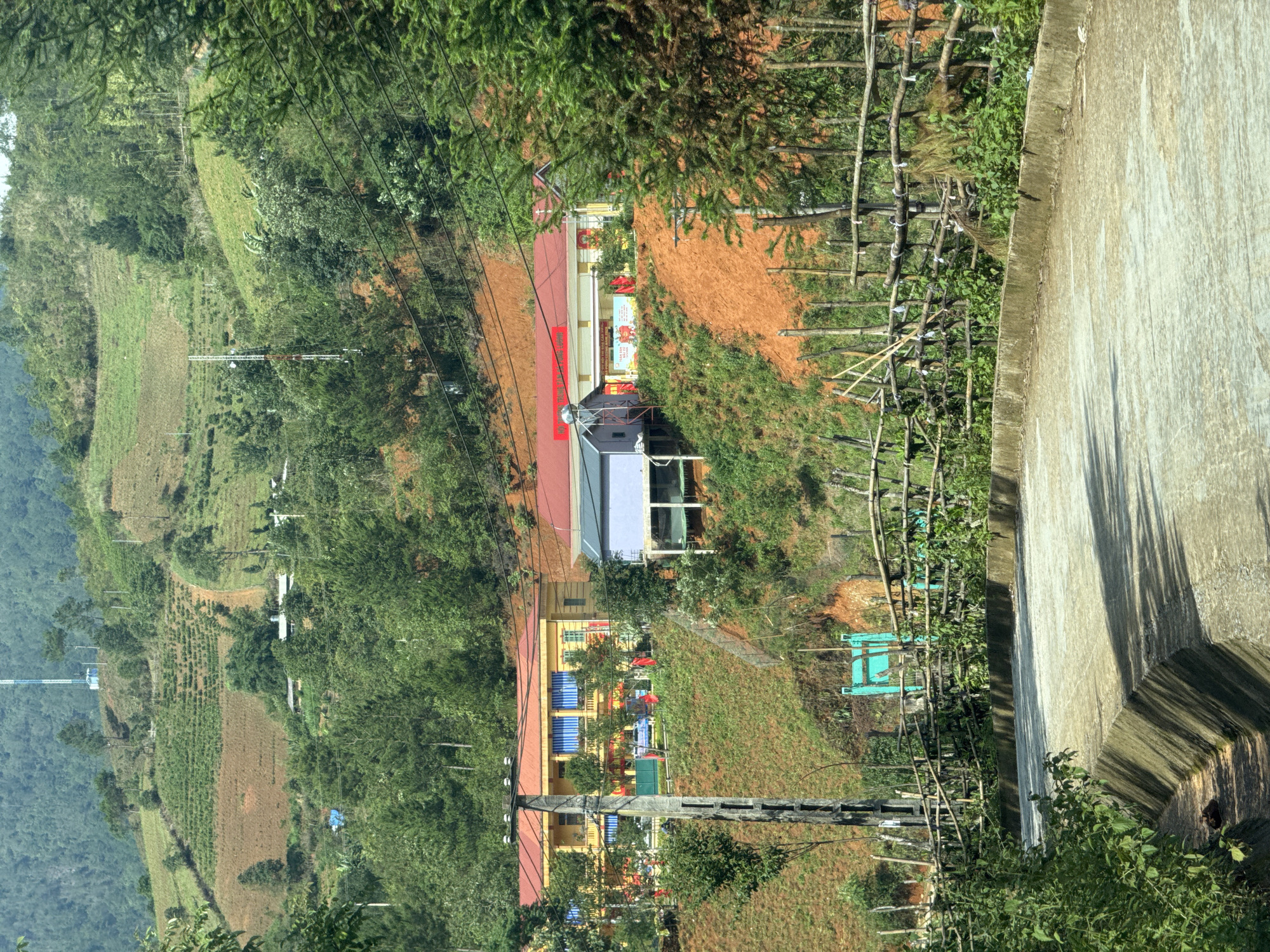 The image depicts a hilly landscape in Púng Luông, Vietnam. Terraced fields are visible, along with scattered vegetation. Buildings, possibly schools or residential structures, are also present. A concrete barrier or wall runs along one edge of the frame. Utility poles and electrical lines crisscross the scene. The photograph appears to have been taken from an elevated position.