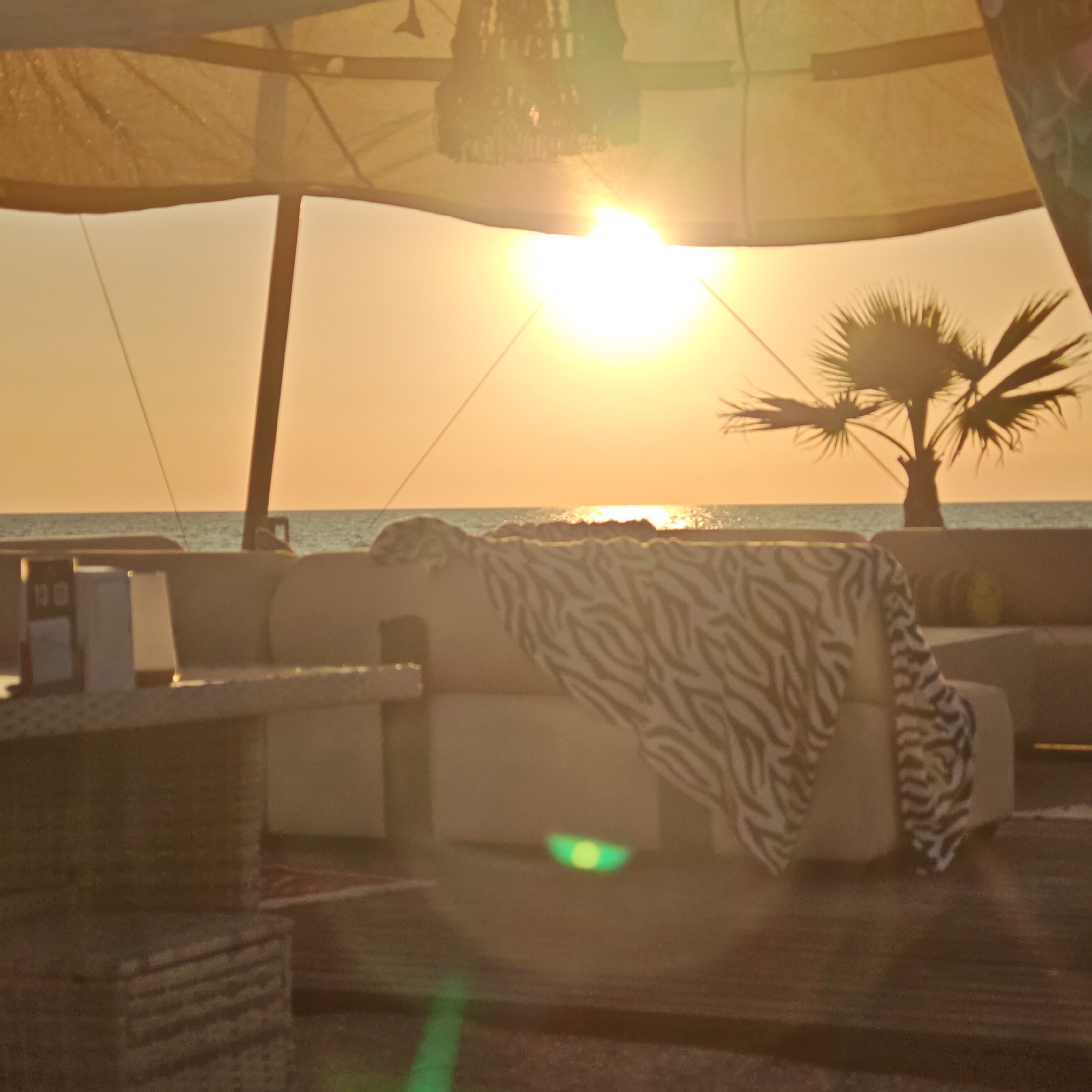 The image depicts an outdoor lounge area set against a coastal sunset. In the foreground, a textured rug is visible, with two woven cube-shaped ottomans or side tables stacked on the left. A light-colored sofa with a zebra-patterned throw blanket draped over it occupies the mid-ground. Behind the sofa, the calm sea meets the horizon under a warm, orange-hued sky illuminated by the setting sun. A silhouette of a palm tree stands to the right of the frame. The lounge appears to be set up under a light-colored canopy, with visible support poles. The scene evokes a tranquil, relaxing atmosphere. There are no people or animals present in the image. The location is identified as an outdoor lounge area in Shchyolkino, Ukraine.