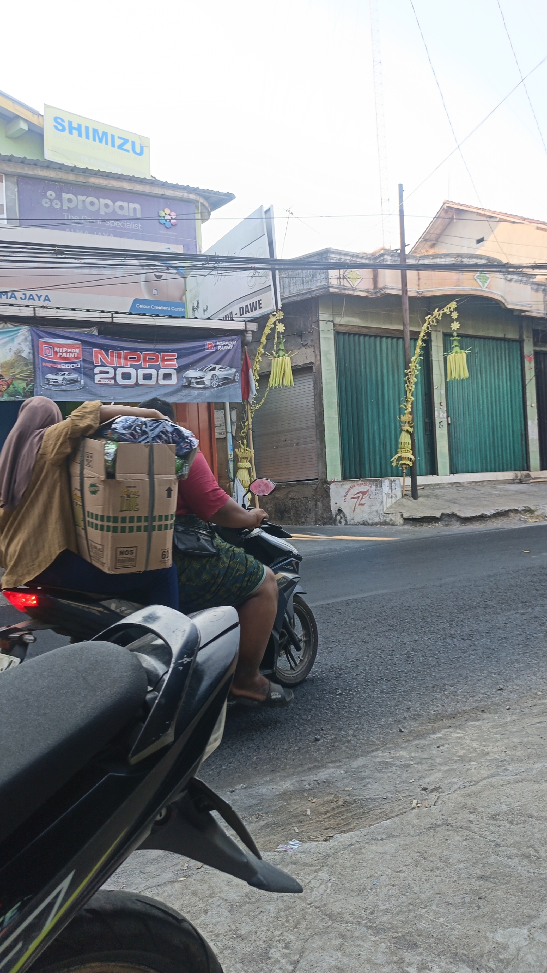 Two individuals are riding a motorcycle on a street in Dawe, Indonesia. The person in the back is wearing a brown hijab and a tan jacket, and they are carrying a large cardboard box strapped to their back. The person in the front is wearing a red shirt and a green patterned skirt, and they are driving the motorcycle. The street appears to be lined with buildings, some of which have green metal roll-up doors. There are banners and signs visible on the buildings, including one that reads "SHIMIZU" and another with "NIPPON PAINT". The sky is bright, suggesting it is daytime. The environment suggests a commercial or residential street in a town or city.