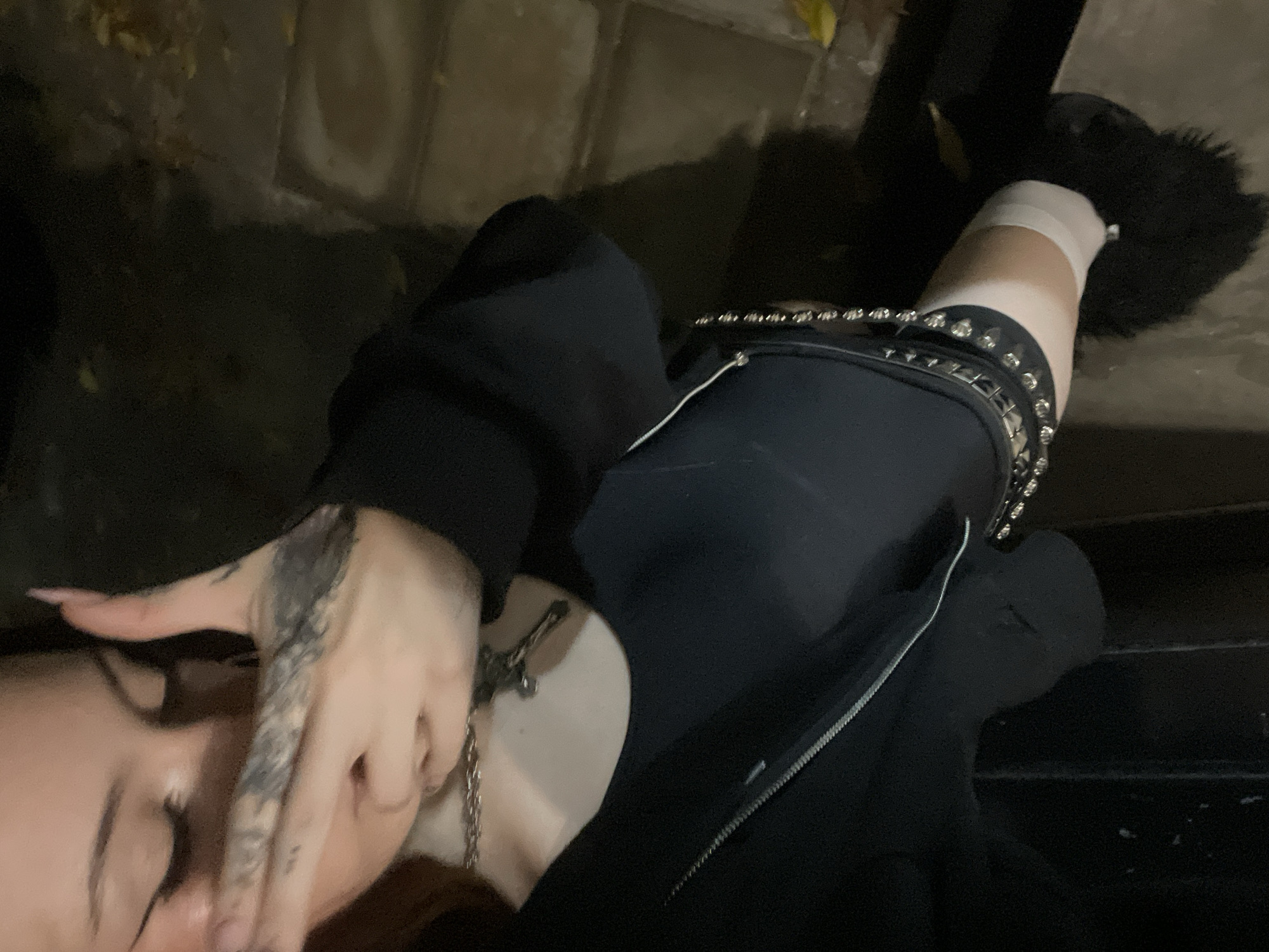 A photograph shows a person sitting on a staircase outdoors, at night. The person has fair skin, tattoos on their hand, and is wearing a silver chain necklace. They are also wearing a black hooded sweatshirt with a silver zipper and a black skirt with a studded belt. Their legs are clad in white socks and black, furry shoes. The background appears to be a stone or concrete walkway or wall. The location in the city of Buenos Aires, Argentina, is not determined.