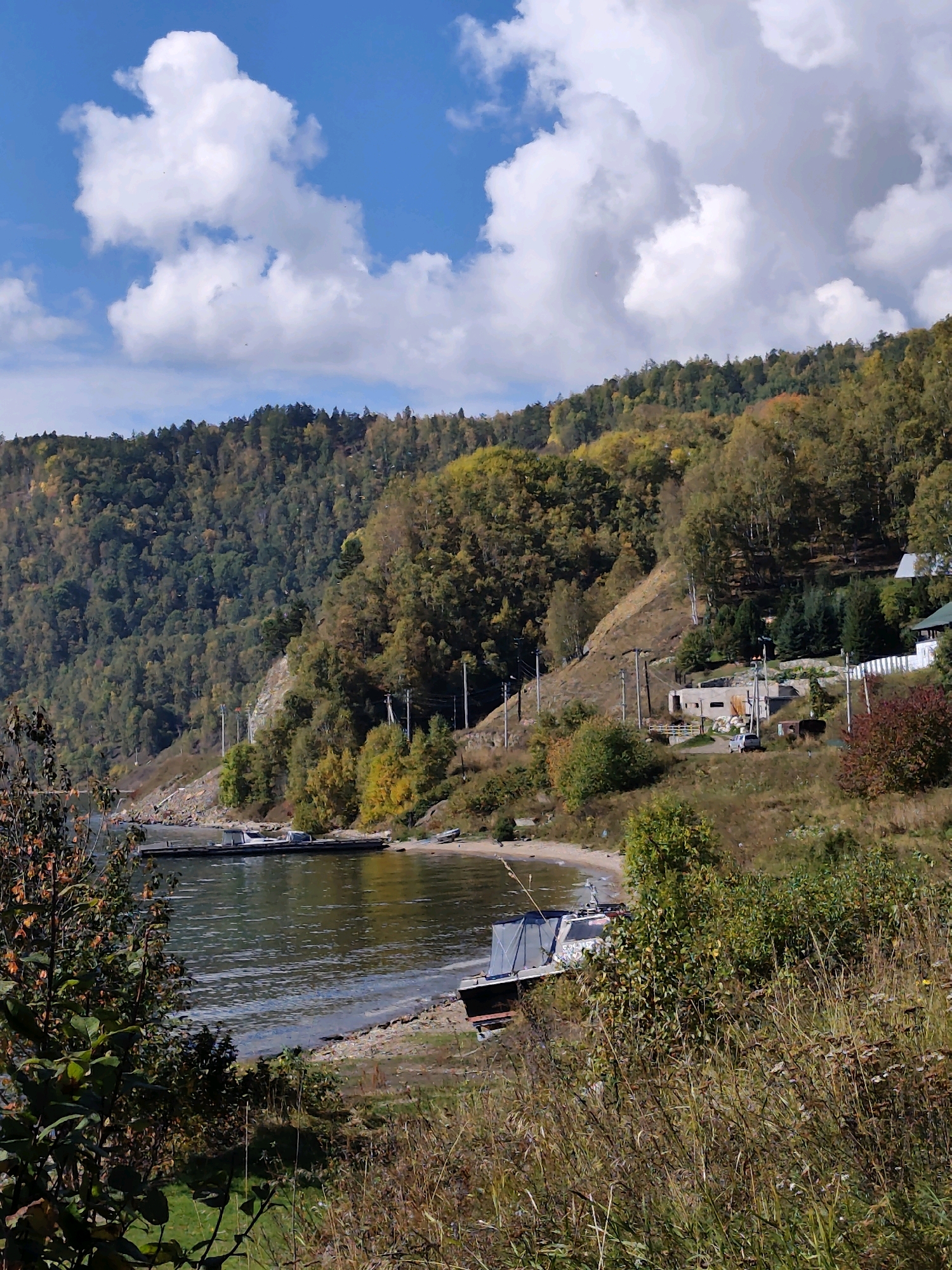 This image depicts a scenic view of a bay on Lake Baikal, Russia, during the autumn season. The foreground is dominated by tall, dry grasses and scattered green bushes, with a few trees on the left showing hints of fall foliage. The water of the bay is calm and reflects the blue sky and white clouds. On the shore, a small, light-colored boat is moored, and further out in the water, a larger, darker vessel is visible.
The background features a steep, tree-covered hillside that stretches across the frame. The trees display a mix of green, yellow, and orange leaves, characteristic of autumn. Along the slope, there are a few buildings and structures, including what appears to be a small, unfinished concrete building with several openings. Power lines and poles are also visible along the hillside. The sky is a bright blue, dotted with large, fluffy white clouds, suggesting a clear and pleasant day. The overall impression is one of natural beauty and a remote, possibly rural, setting along the vast expanse of Lake Baikal.