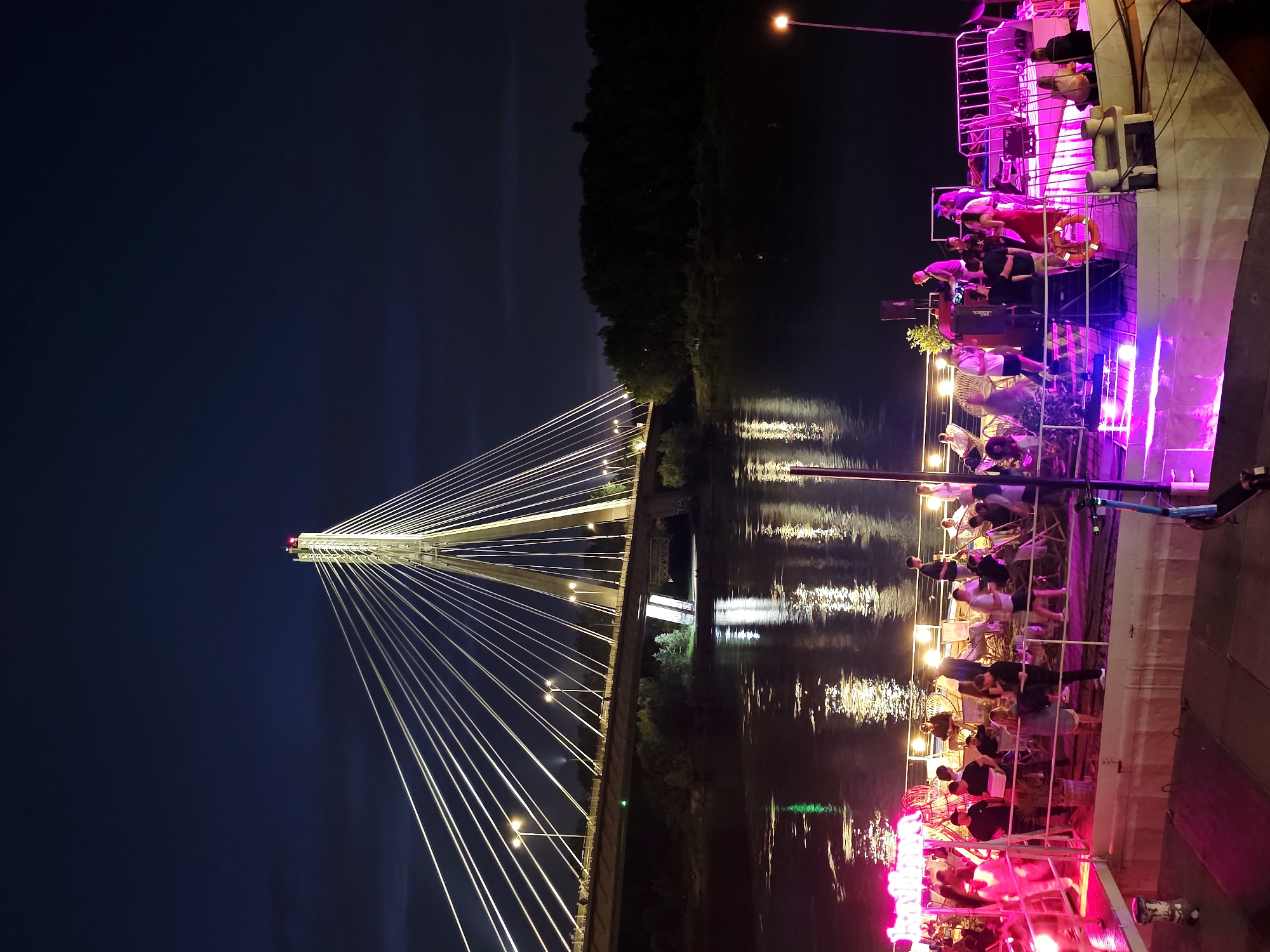This is a nighttime scene in Warsaw, Poland, featuring a lively gathering on a boat docked on the Vistula River, with the Swiętokrzyski Bridge in the background. The bridge, illuminated with bright white lights, dominates the upper portion of the image. Its cable-stayed design is prominent, with numerous cables fanning out from the central tower, which is topped with a red light. The bridge spans across the dark water, its reflection shimmering on the surface.
On the river's edge, the boat is illuminated with vibrant pink and purple lights, creating a festive atmosphere. A crowd of people are visible on the boat, engaged in various activities. Some are standing and conversing, while others are seated in chairs. A DJ booth with speakers is set up on the boat, suggesting music is being played, and a few individuals appear to be dancing. Several scooters are parked on the boat. The dark water of the Vistula River surrounds the boat, with the lights of the city and the bridge reflecting on its surface. Dense trees line the far bank of the river, silhouetted against the night sky.