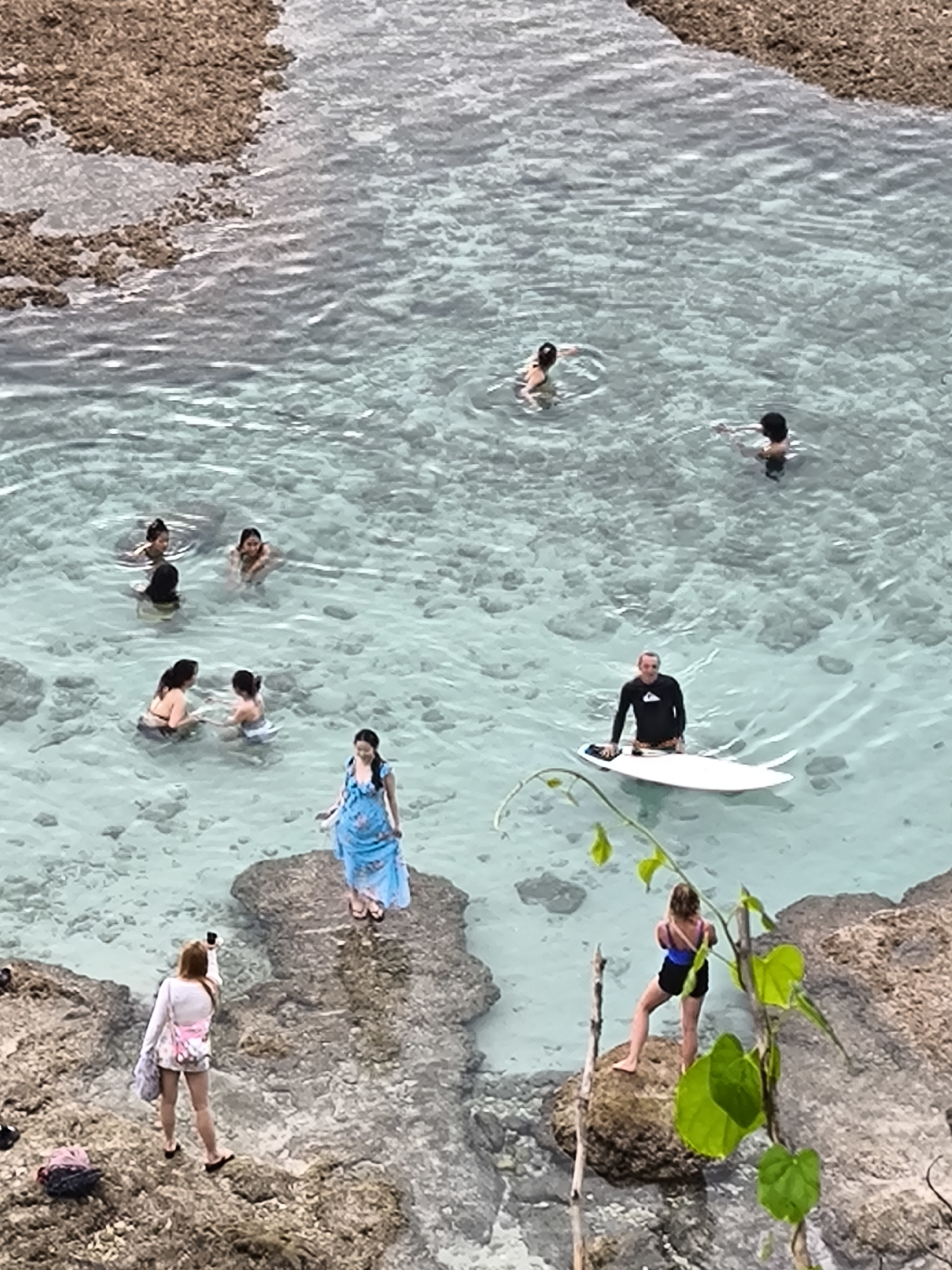 This high-angle shot depicts a clear, shallow cove or natural pool in Uluwatu, Indonesia. Seven individuals are swimming or wading in the water, while another man stands on a white surfboard. On rocky outcroppings surrounding the pool, one woman in a blue dress stands on a rock; another woman takes a photo with a phone; a third woman in a blue and black swimsuit stands on a rock looking out. Vegetation hangs over the edge of the rock. The surrounding landscape is rocky, and some of it is submerged.