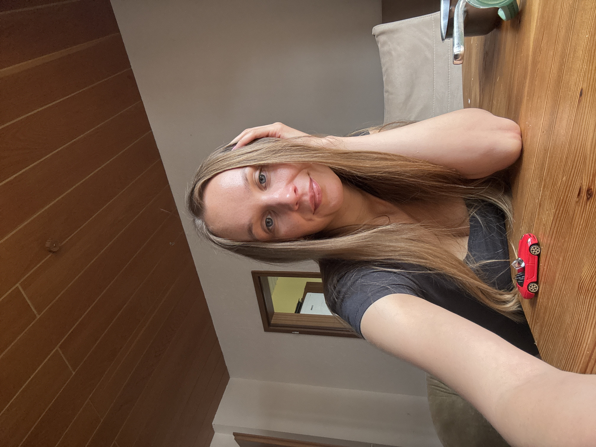 A woman with long, blonde hair and blue eyes is sitting at a wooden table, taking a selfie. She is wearing a dark t-shirt and has her right hand on her head, her arm resting on the table. A small red toy car is on the table in front of her. In the background, there is a doorway, a chair, and a ceiling with wooden planks. The scene appears to be indoors, possibly in a residential setting. The location is identified as Überherrn, Germany.