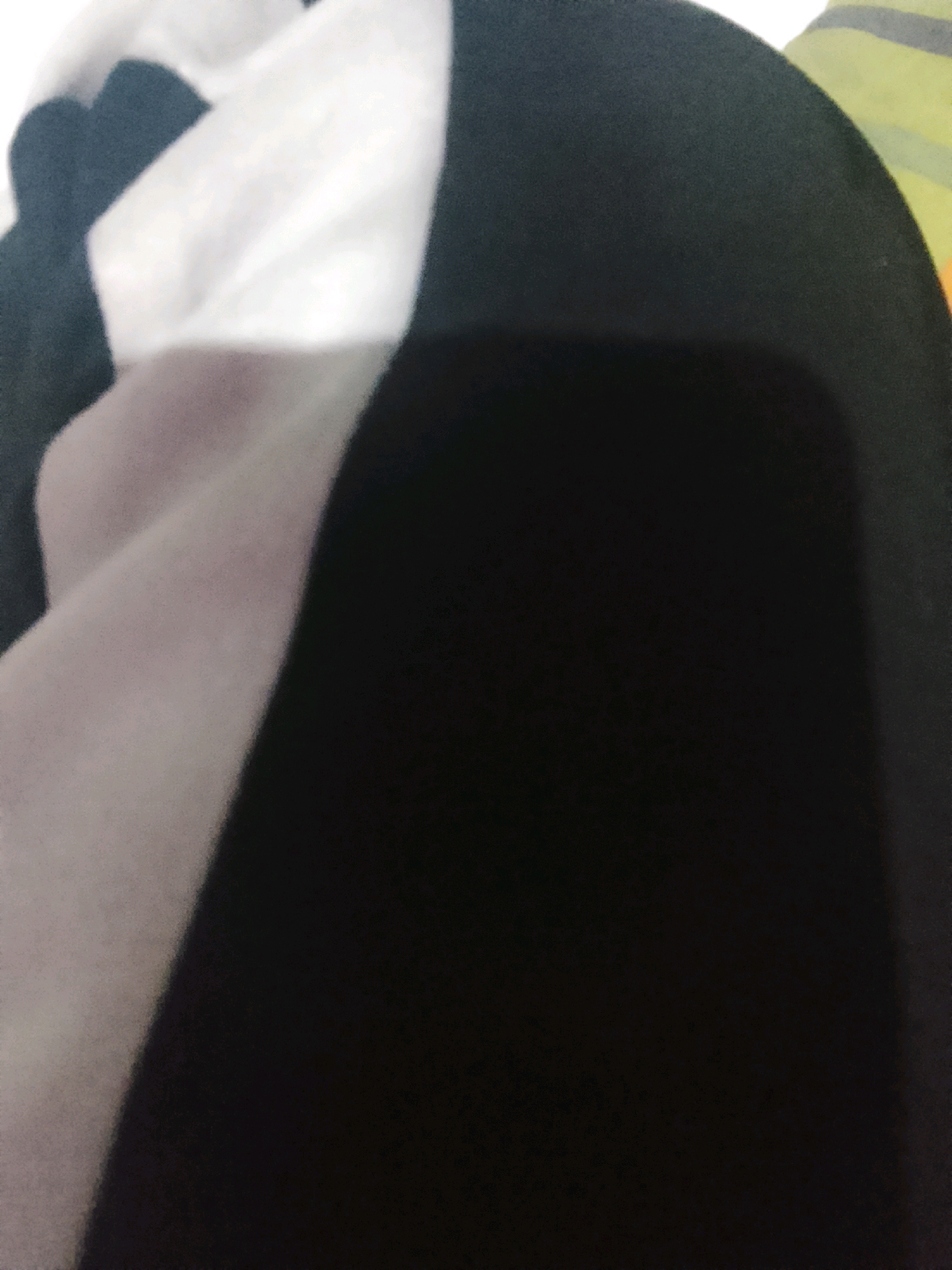 The image shows a close-up of dark fabric with a large, soft shadow cast upon it. The fabric appears to be a part of clothing or a textile. The shadow is not sharply defined, suggesting a diffused light source. The background is out of focus and shows hints of lighter colored fabric with stripes, possibly a colorful garment or bedding. There are no people, specific objects, or contextual environmental elements visible in the image. The location in Sumberwono, Indonesia, is not discernible from the visual information provided in the image.