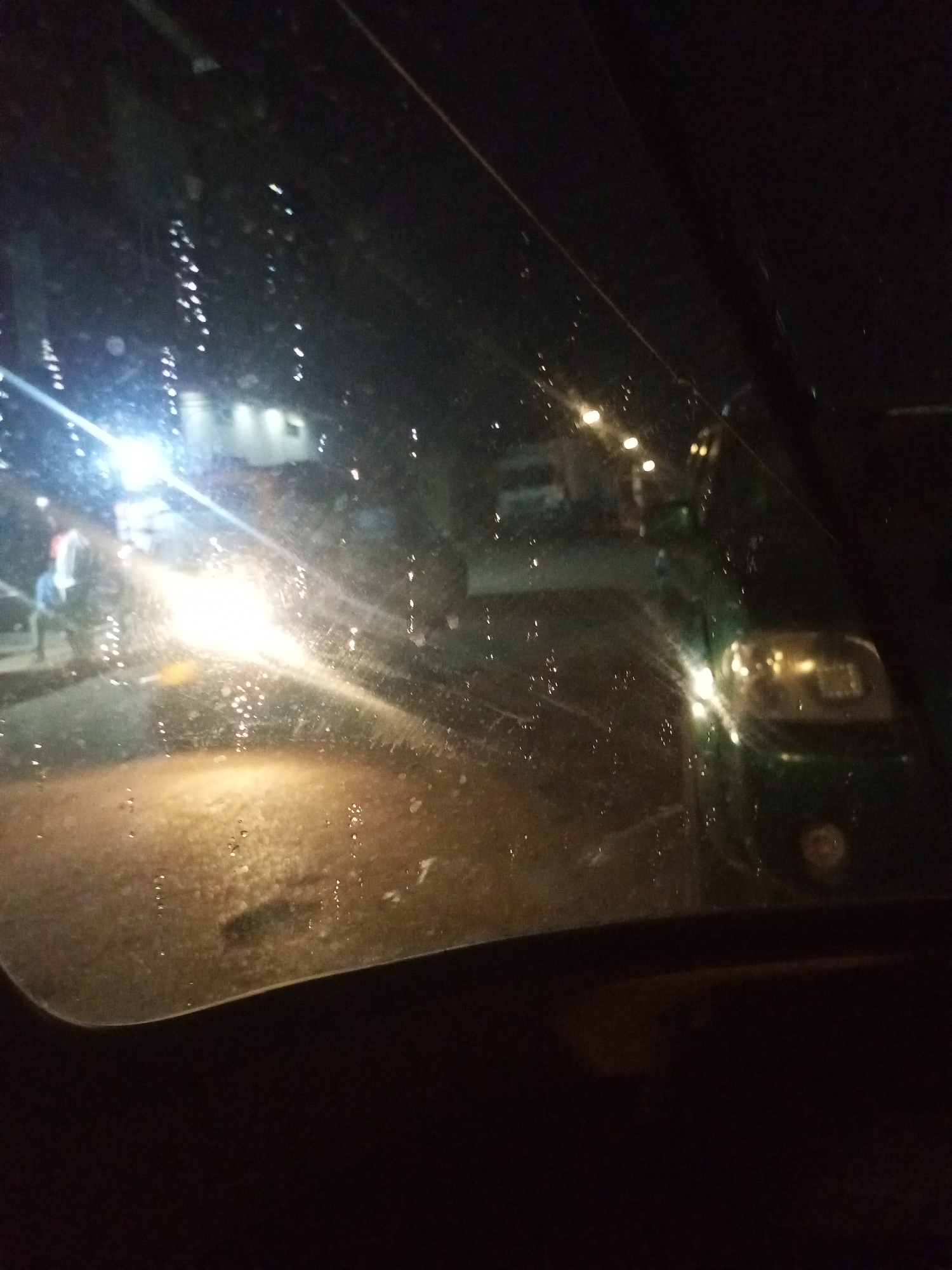 The image captures a nighttime street scene in Maiburiji, Nigeria, viewed from inside a vehicle through a wet or dirty windshield. The viewing surface is covered in numerous water droplets, causing light sources to appear diffused, streaky, and in starburst patterns.
In the left-center of the frame, a powerful, bright light source, likely headlights from an approaching vehicle (possibly a motorcycle or small car), dominates the view, creating a strong glare and obscuring details behind it. To the left of these bright lights, faint, blurred figures that appear to be people are discernible. The road surface in the foreground and mid-ground is reflective and appears wet, consistent with rain or recent rainfall.
On the right side of the image, the front portion of a green vehicle is visible, with its headlights or taillights casting a soft glow. In the background, several diffused artificial lights, possibly from streetlights or buildings, are scattered across the dark landscape. The upper section of the image is largely dark, with subtle traces of ambient light. The bottom portion of the image reveals the dark interior of the vehicle from which the photograph was taken, showing the dashboard or a similar surface.
The scene conveys the experience of navigating a street at night, likely under rainy or recently rainy conditions, as indicated by the wet surfaces and light distortions. No specific text is visible in the image.