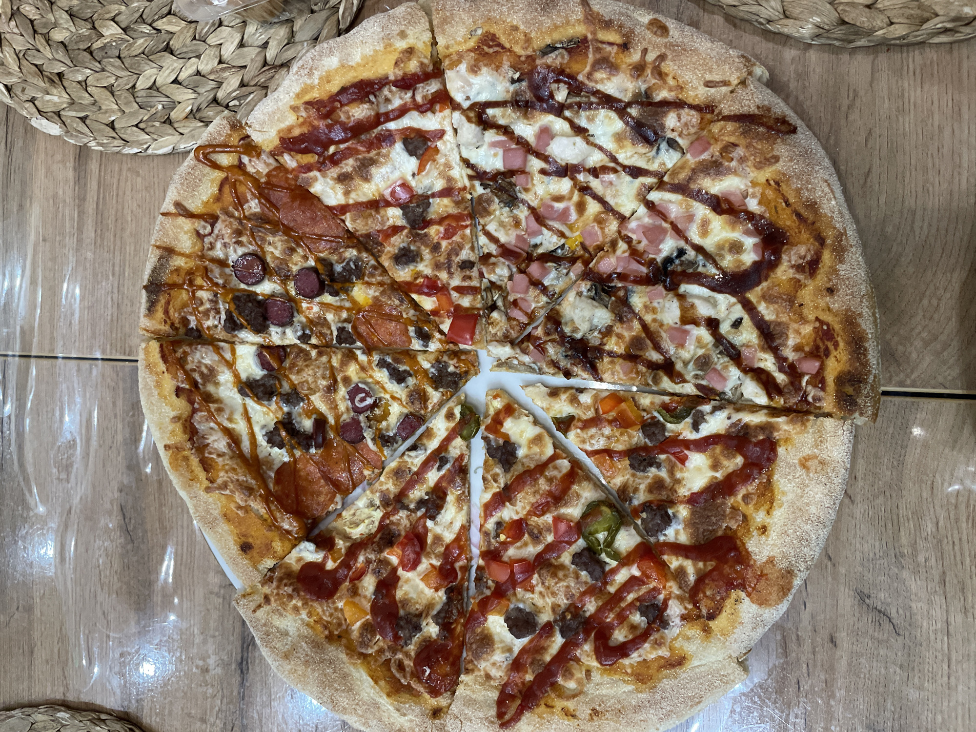 The image shows a sliced pizza with multiple toppings on a wooden surface. The pizza appears to have been cut into several uneven slices. Two place mats are partially visible around the pizza. The location is Лесной Пейзаж Кп (шарапово), Russia. No people are visible.