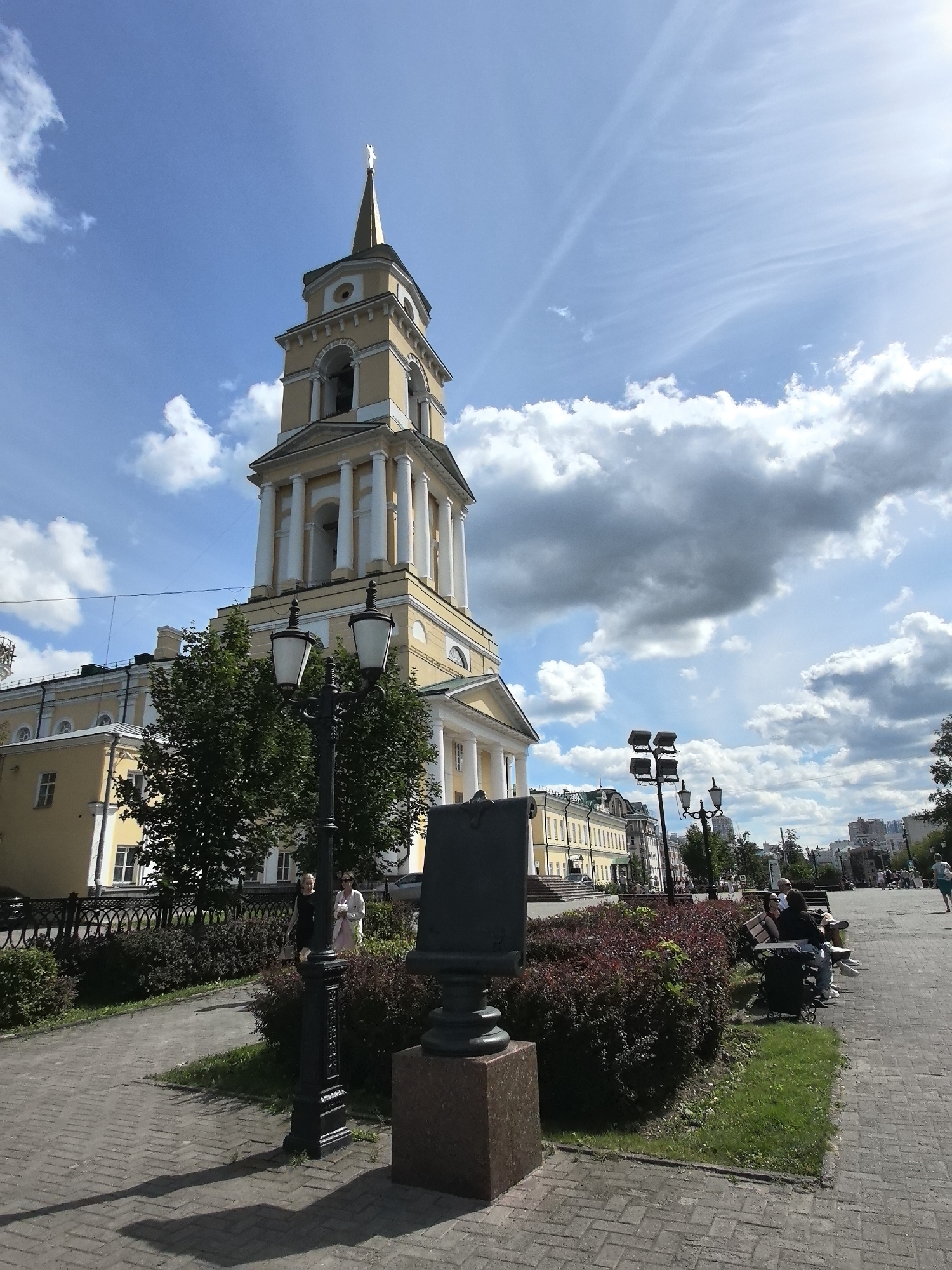 This image captures a sunny day with scattered clouds in front of a large, pale yellow building that appears to be a church or a public institution, with a tall, slender bell tower topped with a cross. The bell tower features classical architectural elements such as columns and arches. In the foreground, a paved walkway runs alongside a garden bed filled with dense, dark red bushes. A black lamppost with two lamps stands prominently on the walkway, casting a long shadow on the brick-patterned pavement. Further down the walkway, several people are seated on benches, some engaged in conversation, while others appear to be resting. A black, rectangular monument or sculpture is positioned to the left of the lamppost. Beyond the main building, other structures and trees are visible, suggesting an urban environment. The overall scene conveys a peaceful, public outdoor space. Based on the architectural style and context, this location is likely the Holy Trinity Cathedral in Perm, Russia.