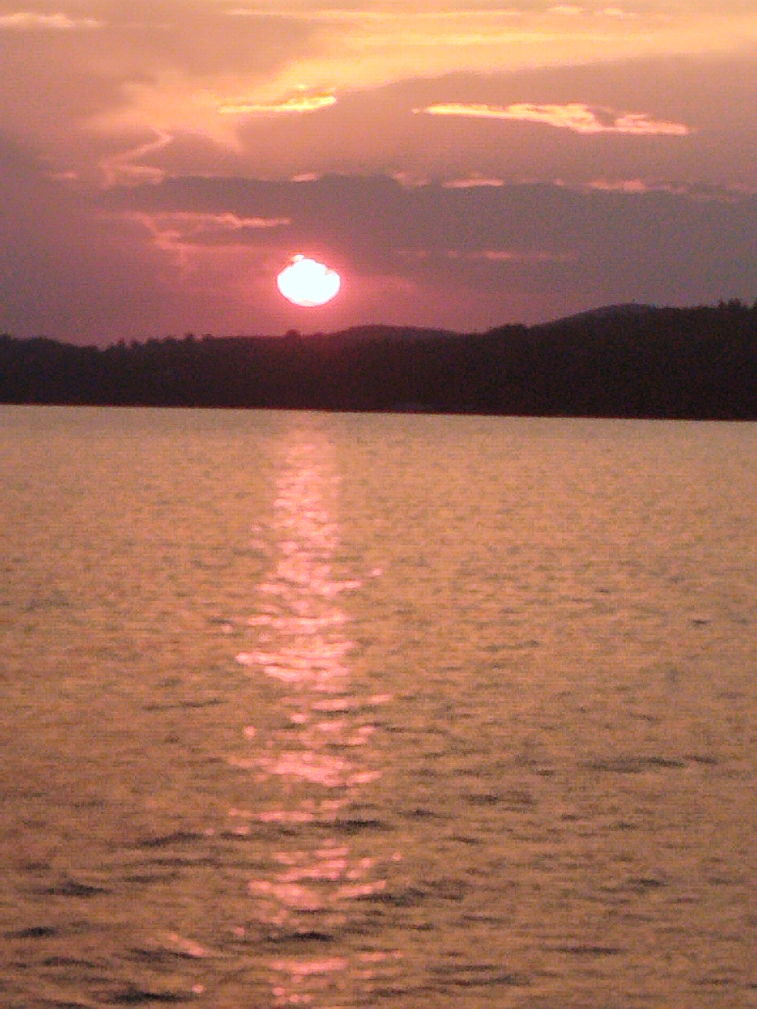 This is a photograph of a sunset over a body of water. The sun is a bright, luminous orb near the horizon, casting a warm, orange glow across the sky and reflecting on the rippling surface of the water. Silhouetted against the sky is a dark, undulating line of trees and hills, likely a shoreline. The overall impression is one of tranquility and natural beauty. The image does not contain any people, specific objects, or actions to describe. The location is not provided in the image.