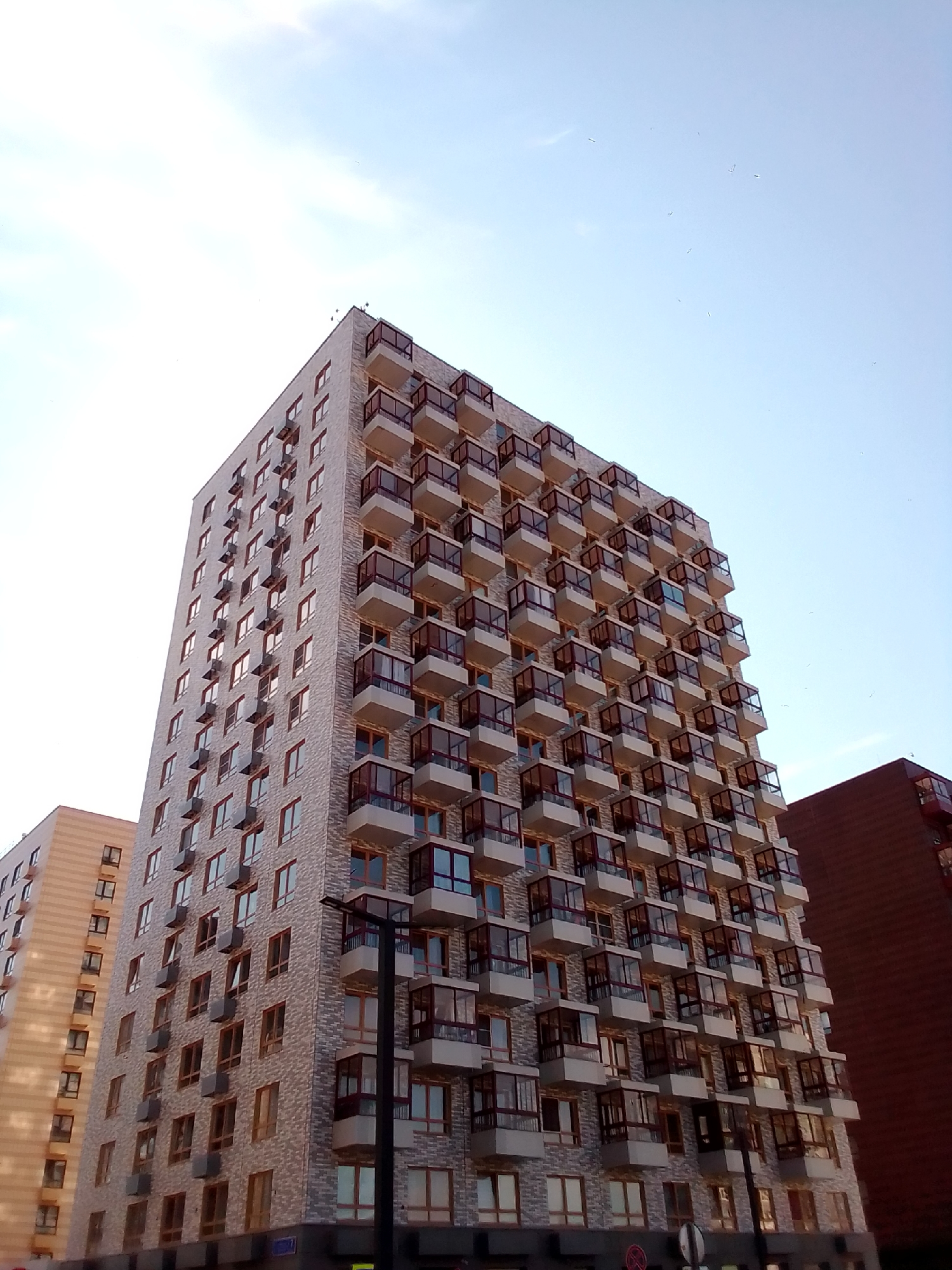 A tall, modern apartment building with numerous balconies is shown from a low-angle perspective against a clear, pale blue sky with scattered clouds. The building's facade is a light, textured material with dark window frames and red-tinted glass on the balconies. Another, darker building is partially visible to the right. Several small dark specks, possibly birds, are scattered across the sky. The image appears to have been taken during daylight hours, with the sky suggesting either early morning or late afternoon.