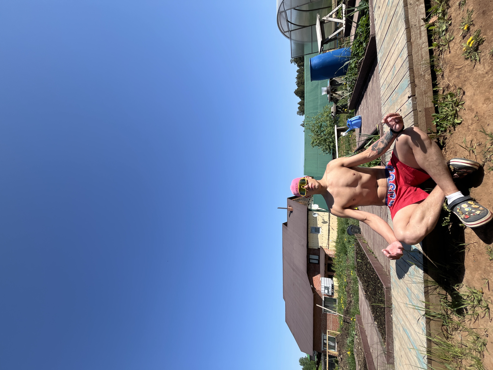 A shirtless man is sitting cross-legged on a wooden boardwalk outdoors, with a clear blue sky above. The man wears red shorts, pink sunglasses, and a pink hat, and has a tattoo on his left arm. He also wears black Crocs with charms and white socks. He is positioned on the right side of the frame. In the background, there's a mix of greenery and buildings. A long green building is visible in the middle ground and behind the man, a house with a brown roof and yellow siding is also present. To the left of the green building, a transparent greenhouse structure can be seen. The location is in a rural area, likely in the vicinity of the city of Chaikovsky, Russia.