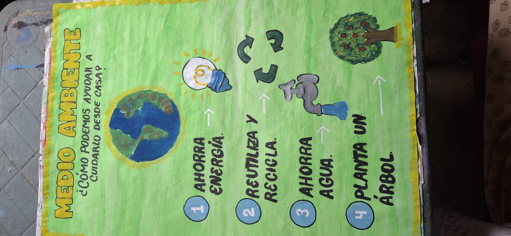 The image depicts a handmade poster with the title "MEDIO AMBIENTE" (Environment). It asks "¿CÓMO PODEMOS AYUDAR A CUIDARLO DESDE CASA?" (How can we help take care of it from home?). The poster presents four actions: 1) AHORRA ENERGIA (Save Energy) with a picture of the earth and a light bulb; 2) REUTILIZA Y RECICLA (Reuse and Recycle) with the recycling symbol; 3) AHORRA AGUA (Save Water) with a picture of a faucet; and 4) PLANTA UN ÁRBOL (Plant a Tree) with a picture of a tree. The poster is painted with bright colors, primarily green, and is presumably located in Vallecito, Venezuela.