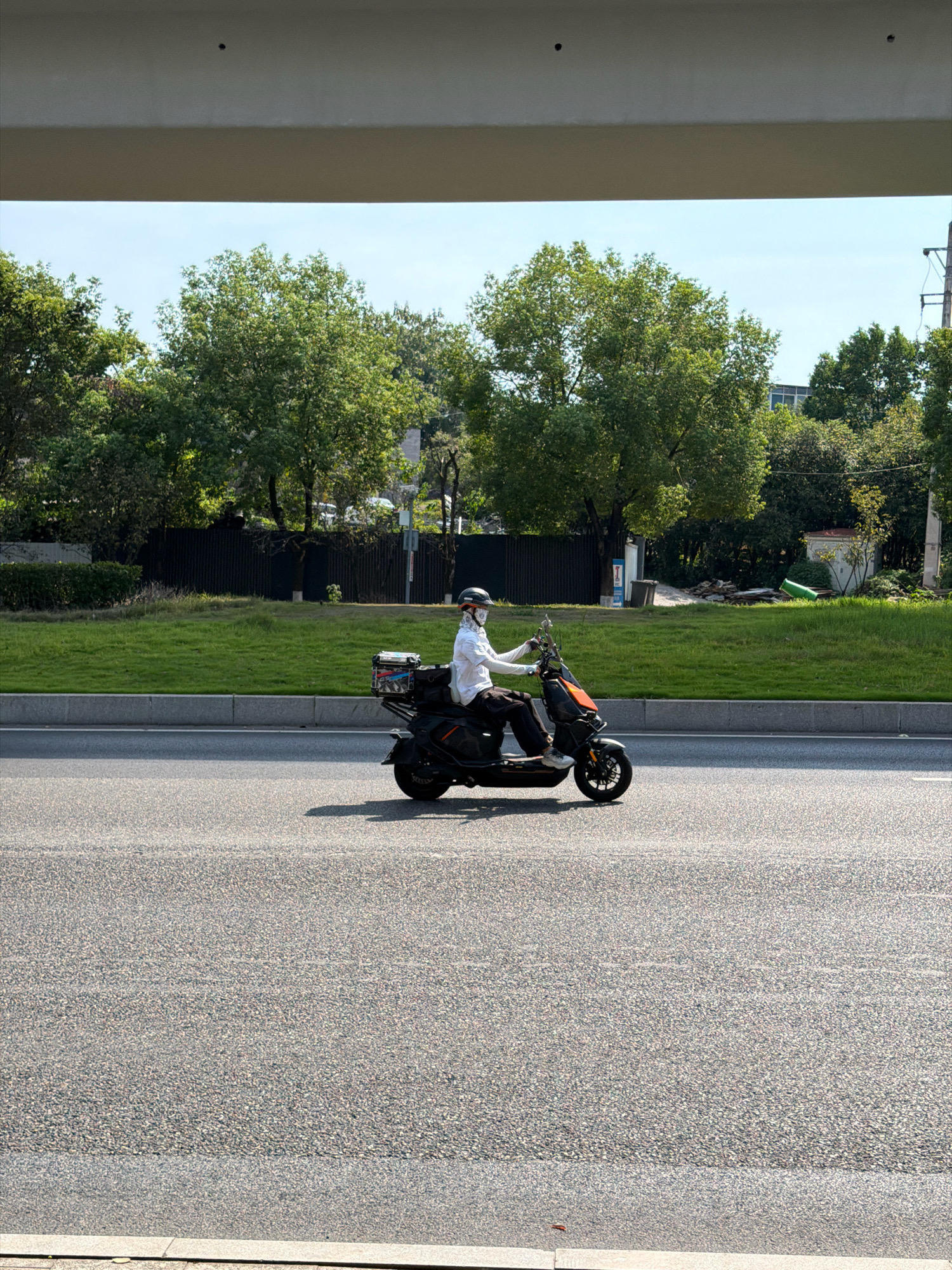 A person rides a black electric scooter on a paved road in Jiangjin, China. The person wears a white long-sleeved shirt, dark pants, a helmet, and a patterned face covering. The scooter has a cargo box on the back, which appears to contain several items. The road is divided into lanes by white lines. To the left of the road is a grassy median with a curb. Beyond the median, there are trees and a dark fence. In the distance, there are buildings and utility poles. The sky is clear and blue. The image is taken from a slightly elevated perspective, possibly from an overpass or a building.