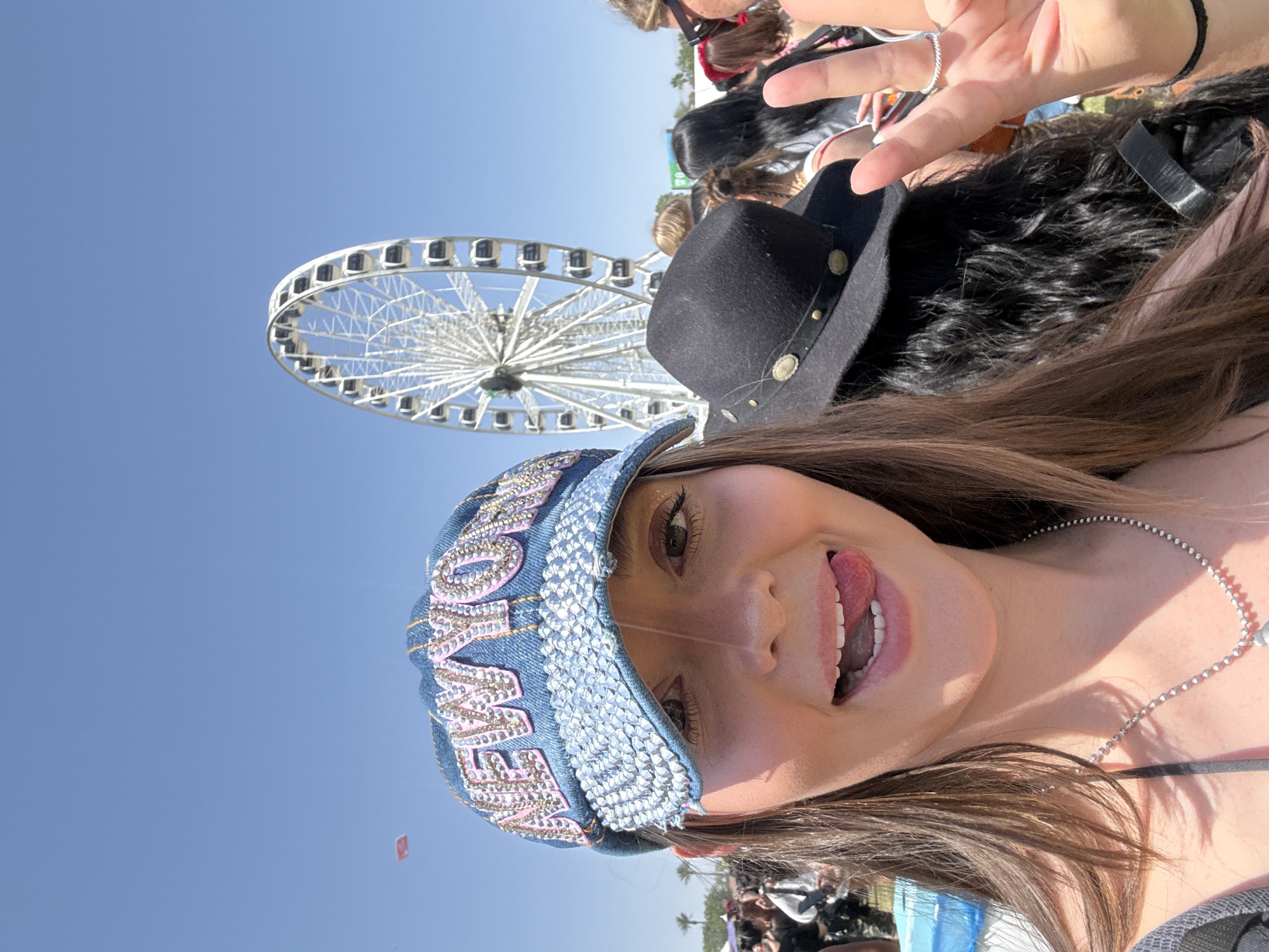 The image is a selfie taken in Indio, California. A young woman with brown hair is the main subject, wearing a denim cap adorned with rhinestones, with the word "COWBOY" embroidered on it. She is sticking her tongue out and making a peace sign with her right hand. Behind her is a Ferris wheel, likely at a festival. A black cowboy hat is positioned near her head, and other people are visible in the background, indicating a crowded outdoor event. The sky is clear and blue.