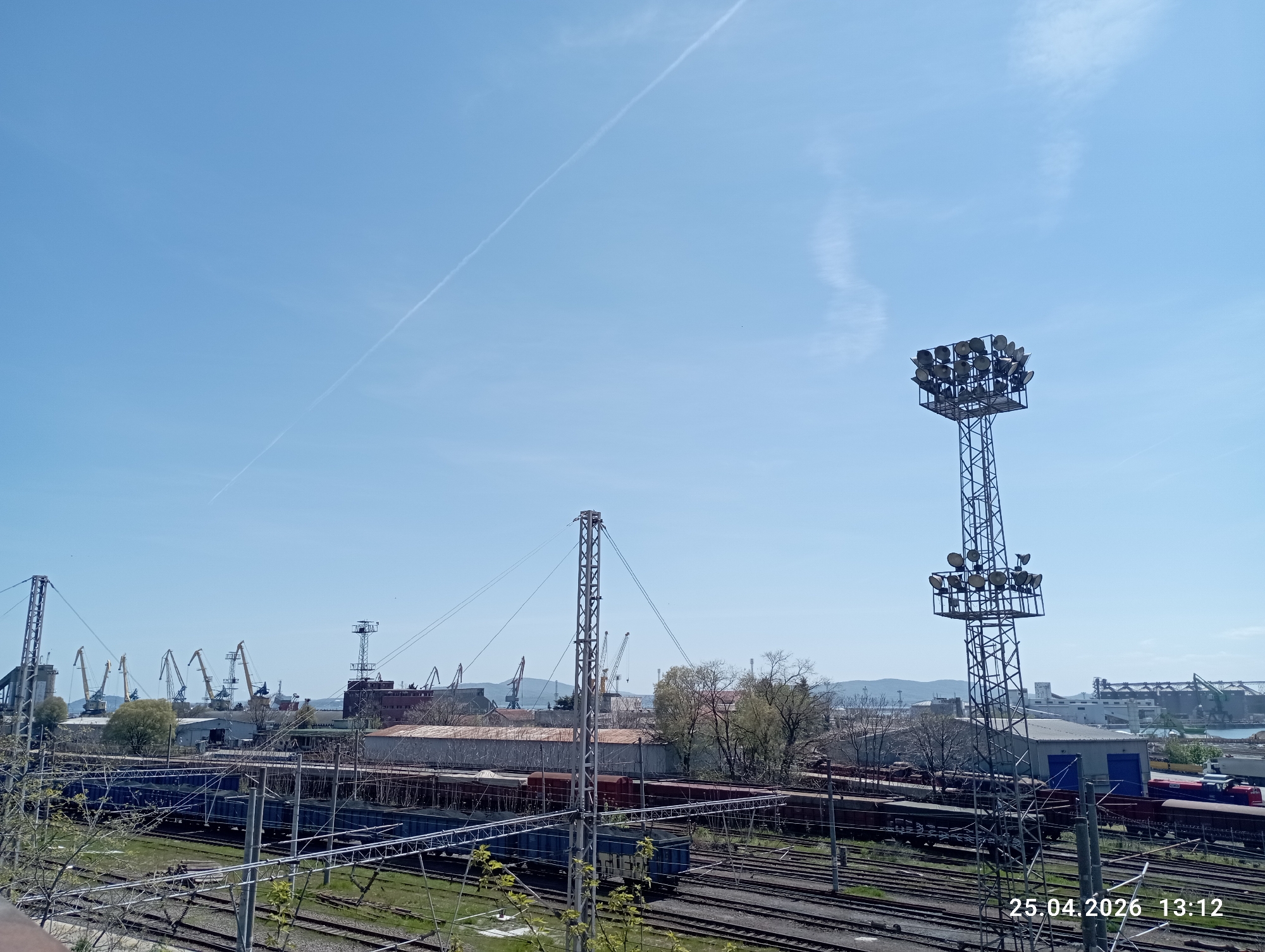 The image depicts a railway yard in an industrial area with several train tracks and train cars present. Two tall metal towers are visible; the taller one is equipped with numerous spotlights. In the background, cranes and buildings indicative of a port or industrial complex can be seen. The sky is clear with a contrail crossing it. There is a date stamp of "25.04.2026 13:12" visible in the bottom right corner of the image.