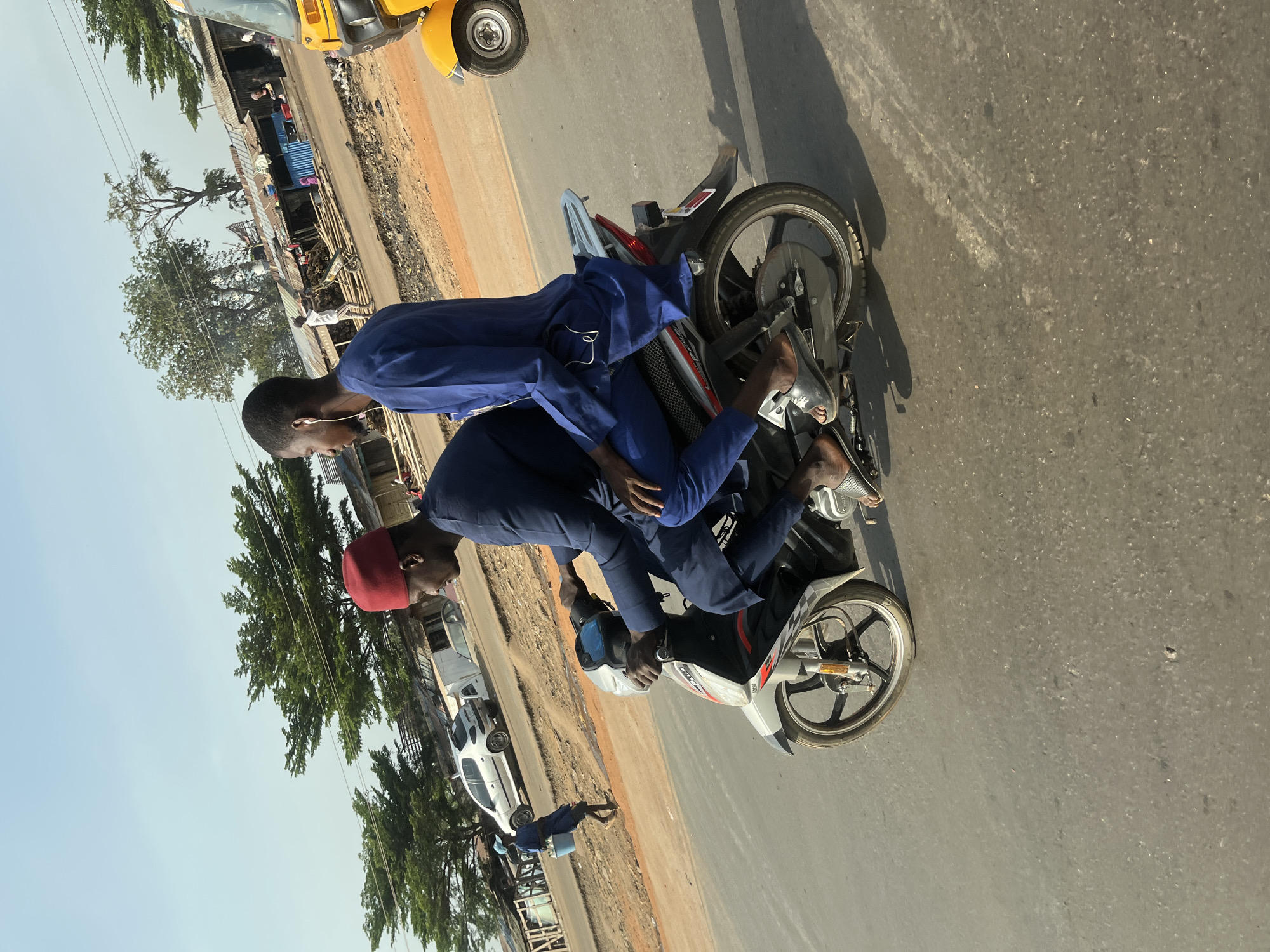 An image, correctly oriented, captures a daytime street scene in Ado, Nigeria. Two men are riding a silver and black motorcycle on an asphalt road. The front rider wears a navy blue outfit and a red cap, with his hands on the handlebars. Behind him, a passenger in a matching navy blue outfit and white wired earphones sits, looking slightly downwards.
To the left of the road, an unpaved shoulder borders a row of simple, low-rise buildings with corrugated iron roofs. Various trees and greenery are interspersed along this side, and some blue containers are visible near the structures. In the background on the left, a yellow Keke Napep (auto-rickshaw) is partially seen, and a white car is parked further down the road. Another individual, dressed in blue and possibly carrying a bucket, walks along the unpaved shoulder.
The clear sky and long shadows indicate either morning or late afternoon. The scene portrays a typical day in an active community. No visible text is clearly legible in the image.