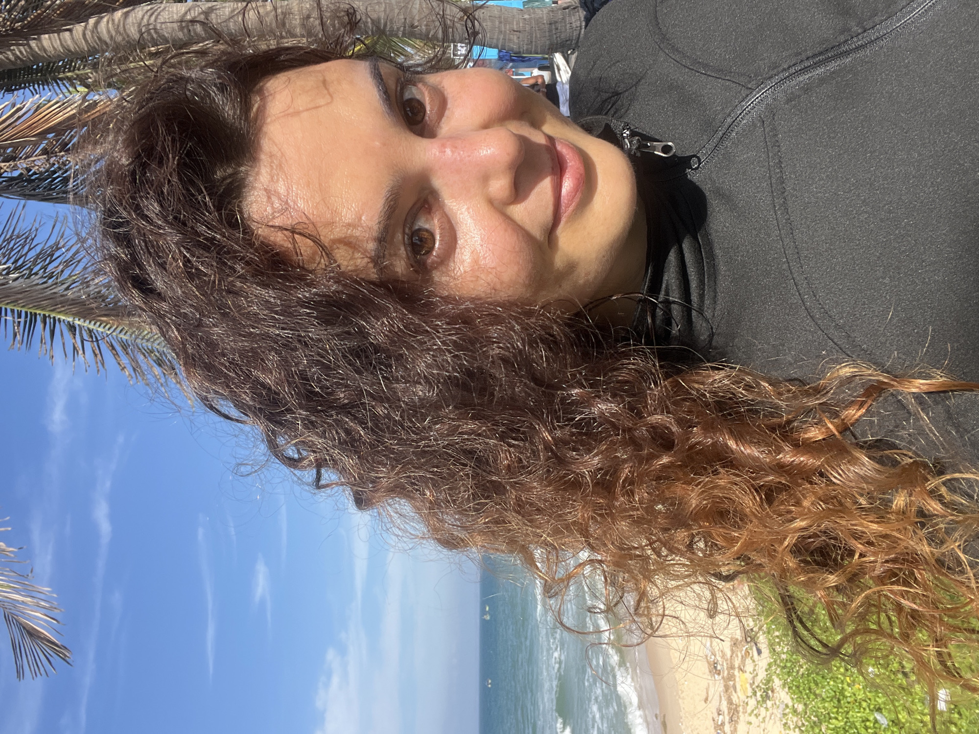 A woman with long, curly brown hair is lying down and looking at the camera. She is wearing a black zip-up jacket. Her face is illuminated by the sun, and she has a relaxed expression. The background shows a beach with turquoise water and a clear blue sky. Palm trees are visible on the left side of the image. The sun appears to be high in the sky, suggesting it is daytime. The overall scene is a casual, outdoor setting, likely a beach vacation or outing. The image is captured in Phu Thuy Ward, Vietnam.