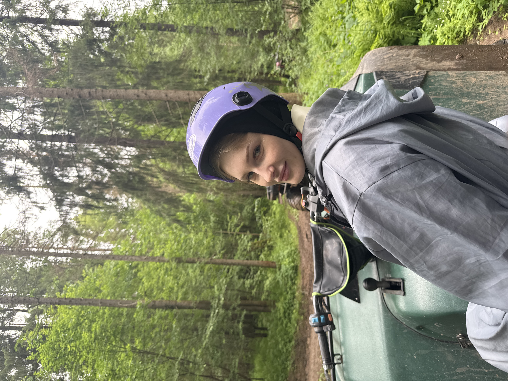 A young adult female is depicted on a green all-terrain vehicle (ATV) in a dense forest environment, specifically located near Perm, Russia. She is positioned with her back partially to the viewer, turning her head over her right shoulder to face the camera directly. She wears a light purple open-face helmet with black ear covers, a black chin strap secured with a red clasp, and a grey/silver hooded jacket, the hood pulled up. Her facial expression is a slight, serene smile, and a faint, light-colored mark is visible on her right cheek below her eye.
The ATV is green and shows signs of being mud-splattered on its front fender and the visible right tire, indicating recent off-road activity. Black handlebars with blue and orange controls are prominent on the left side of the frame, alongside a black storage pouch with a neon green trim attached to the front of the ATV. A black gear shifter lever is visible centrally on the ATV's body, between the handlebars.
The setting is a lush, verdant forest characterized by numerous tall, slender tree trunks and a dense undergrowth of various green foliage, including ferns. The ground beneath the ATV is a dirt track, appearing damp and muddy. The lighting is diffused, suggesting an overcast day or a heavily canopied area, with no harsh shadows present.