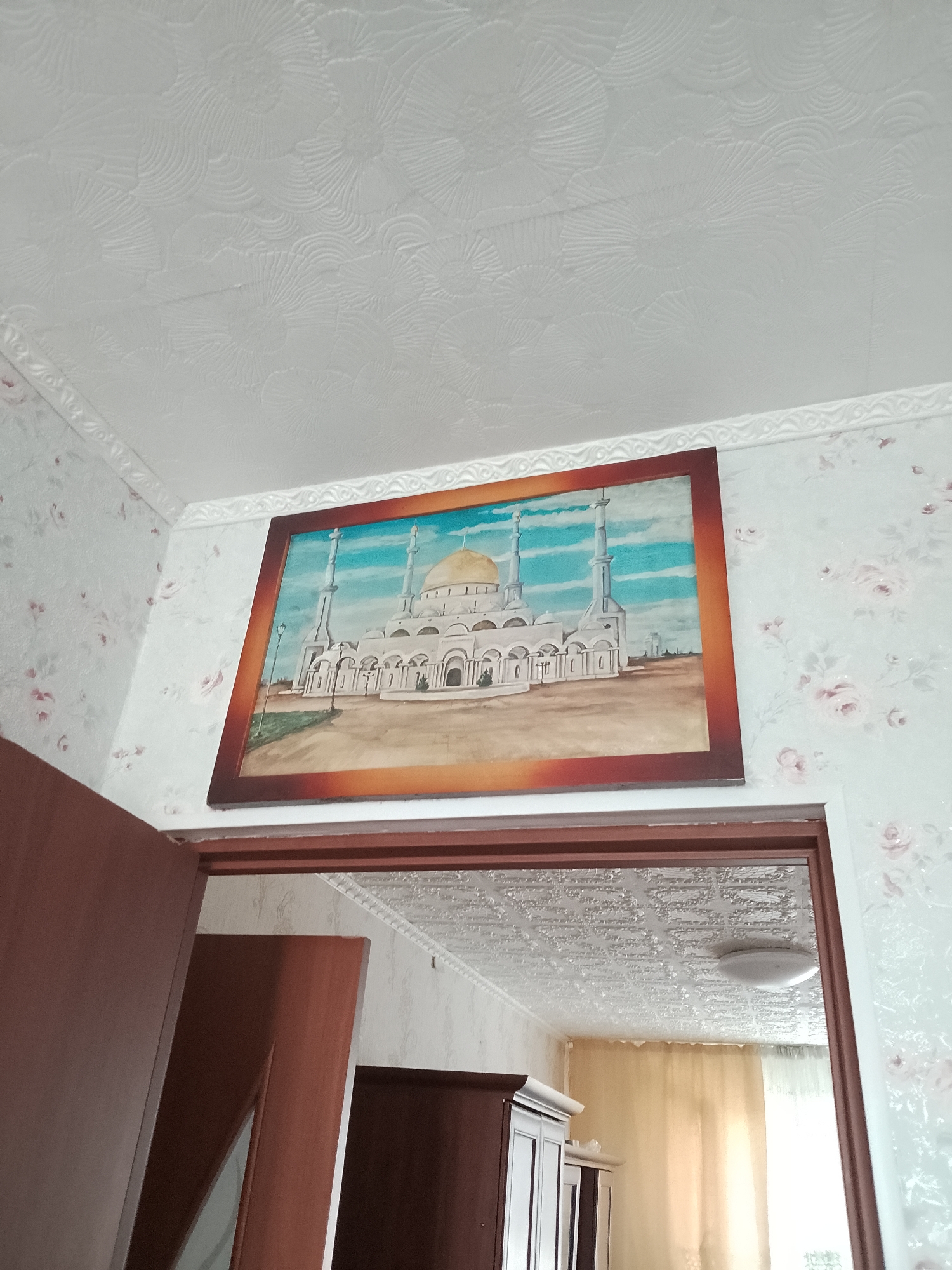 The image shows an interior view of a room in the city of Abay, Kazakhstan. A framed painting of a white mosque with a golden dome and two minarets hangs on a wall. The wall is covered with floral wallpaper and a decorative ceiling border. Below the painting is a doorway leading to another room. The room visible through the doorway has a textured ceiling and curtains. To the left of the doorway is a brown wooden door.