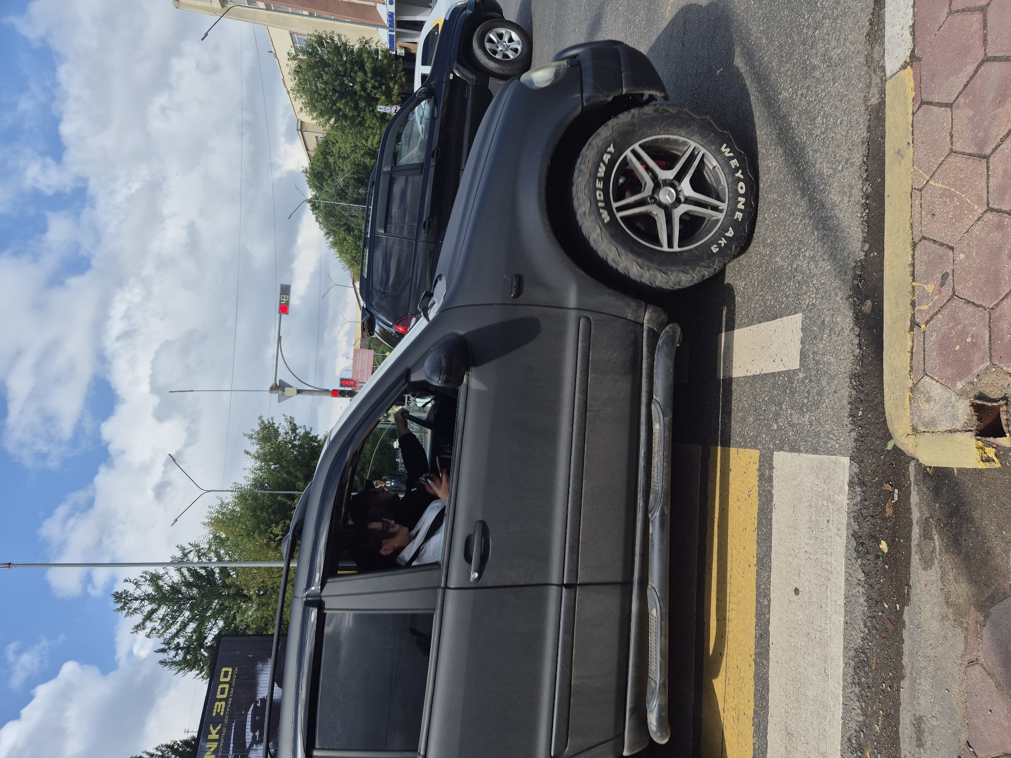 The image shows a black SUV stopped at a pedestrian crossing on a street in Kokshetau, Kazakhstan. The driver, a male, is partially visible behind the steering wheel. A female passenger in the front seat is looking at her phone. The car has a matte black finish and is equipped with a roof rack. In the background, another black SUV is also stopped behind the first. Traffic lights are visible, with a red light illuminated. Trees and buildings line the street, and the sky is partly cloudy. The pedestrian crossing has white and yellow markings. The tire of the foreground SUV has "WIDEYONE AK3" and "WIDEWAY" written on it.