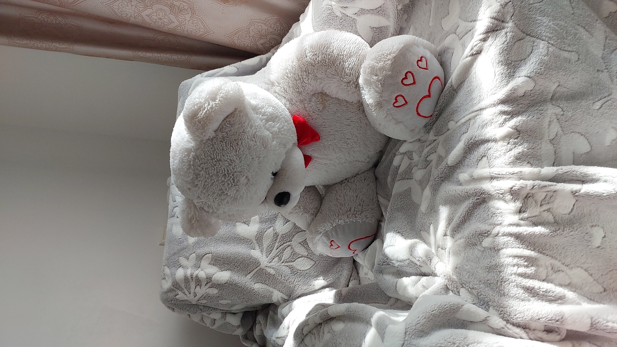 A white, plush teddy bear with a red bow sits on a grey blanket with a floral pattern. The bear is positioned on its right side, with its head bowed. Its paws are adorned with red hearts. The blanket is draped over a chair, and sunlight streams in from the right, casting shadows across the scene. The background consists of a wall and a beige curtain with a patterned design. There is no information in the image to indicate its location in Novocheboksarsk, Russia.