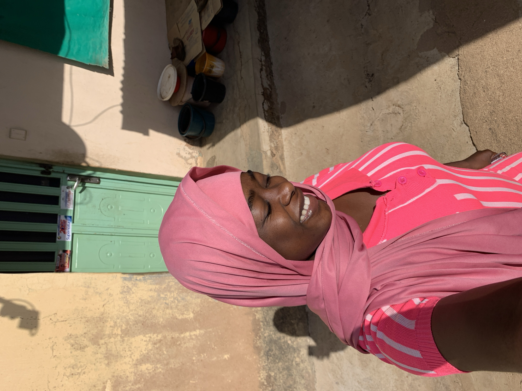 A smiling woman wearing a pink headscarf and a pink and white striped top is standing outdoors in Rafin Par, Nigeria. She is positioned on a concrete surface adjacent to a building with a pale yellow wall. To the left is a green door and above, on the building's wall, is a green patch. Discarded items, including paint buckets, are stacked against the wall next to the door. The lighting suggests a sunny day, casting shadows on the ground.