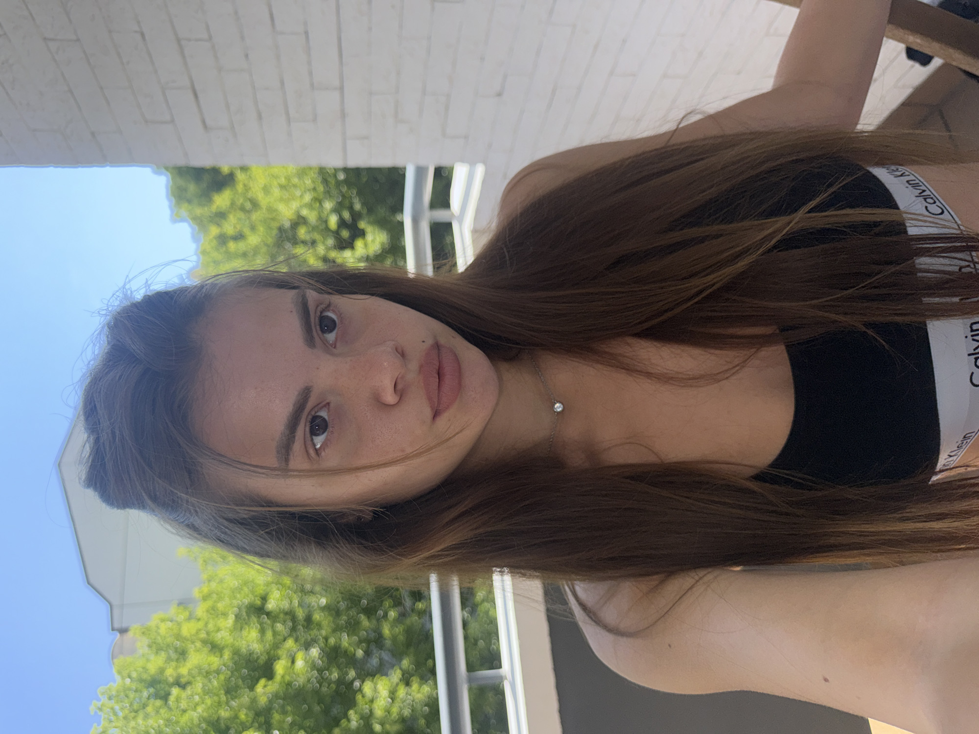 The media file features a close-up, selfie-style portrait of a young woman with long, brown hair. She is wearing a black top with a white band that reads "Calvin Klein." Her head is tilted, and she is looking directly at the camera. Her expression is neutral.
The background consists of a bright blue sky and lush green trees, suggesting an outdoor setting during the daytime. There is also a glimpse of a white brick wall and some structural elements that might indicate a balcony or an outdoor seating area. The lighting suggests it is a sunny day.
The image is oriented vertically, with the subject appearing to be leaning into the frame. The composition is focused on the woman's face and upper body. The overall impression is casual and relaxed, possibly taken during a summer day. The context provided places the image in Plovdiv, Bulgaria.