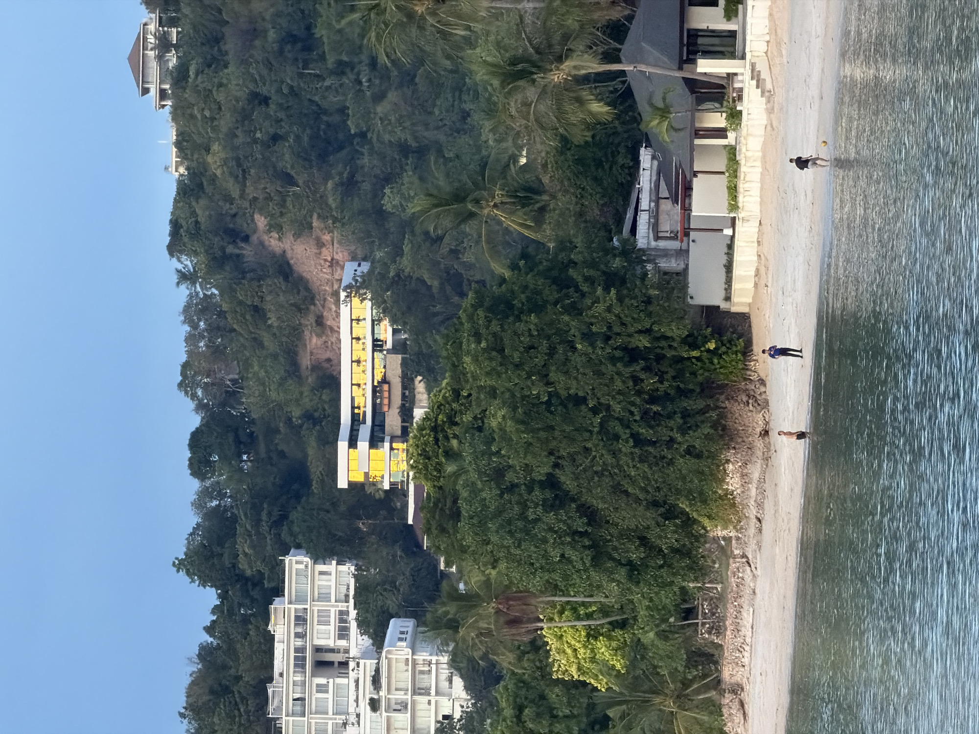 The image captures a waterfront view of Baan Bang Rak, Thailand, during what appears to be late afternoon or early evening, judging by the soft, diffused light. The scene is dominated by a lush, green hillside populated by dense tropical foliage. Several buildings are visible on the hillside. Towards the left, a modern white multi-story building with many windows is perched on the slope. Adjacent to it, further up the hill, is another contemporary building with a distinct yellow facade and numerous windows reflecting the ambient light. Higher up on the crest of the hill, a large, luxurious house with a brown-tiled roof is visible, offering a panoramic view of the surroundings.
In the foreground, a sandy beach stretches along the waterline, with a calm body of water in front of it. The water has a gentle ripple pattern, indicating minimal wave action. Scattered on the beach are a few individuals. On the right side of the beach, two young males are engaged in an activity. One, wearing a dark t-shirt and shorts, is near the water's edge, interacting with a yellow ball. The other, further up the beach, is wearing a blue shirt and dark pants and appears to be observing or waiting. Another person, a male in dark shorts, is standing closer to the water on the left side of the frame.
Behind the beach, a low stone wall separates the sand from the vegetation. A modern, single-story building with a dark, sloped roof and white walls is situated on the right side of the beach, featuring an open-air patio area with pillars. Palm trees are interspersed throughout the landscape, contributing to the tropical ambiance. The overall scene suggests a serene coastal environment with a blend of natural beauty and modern architecture.