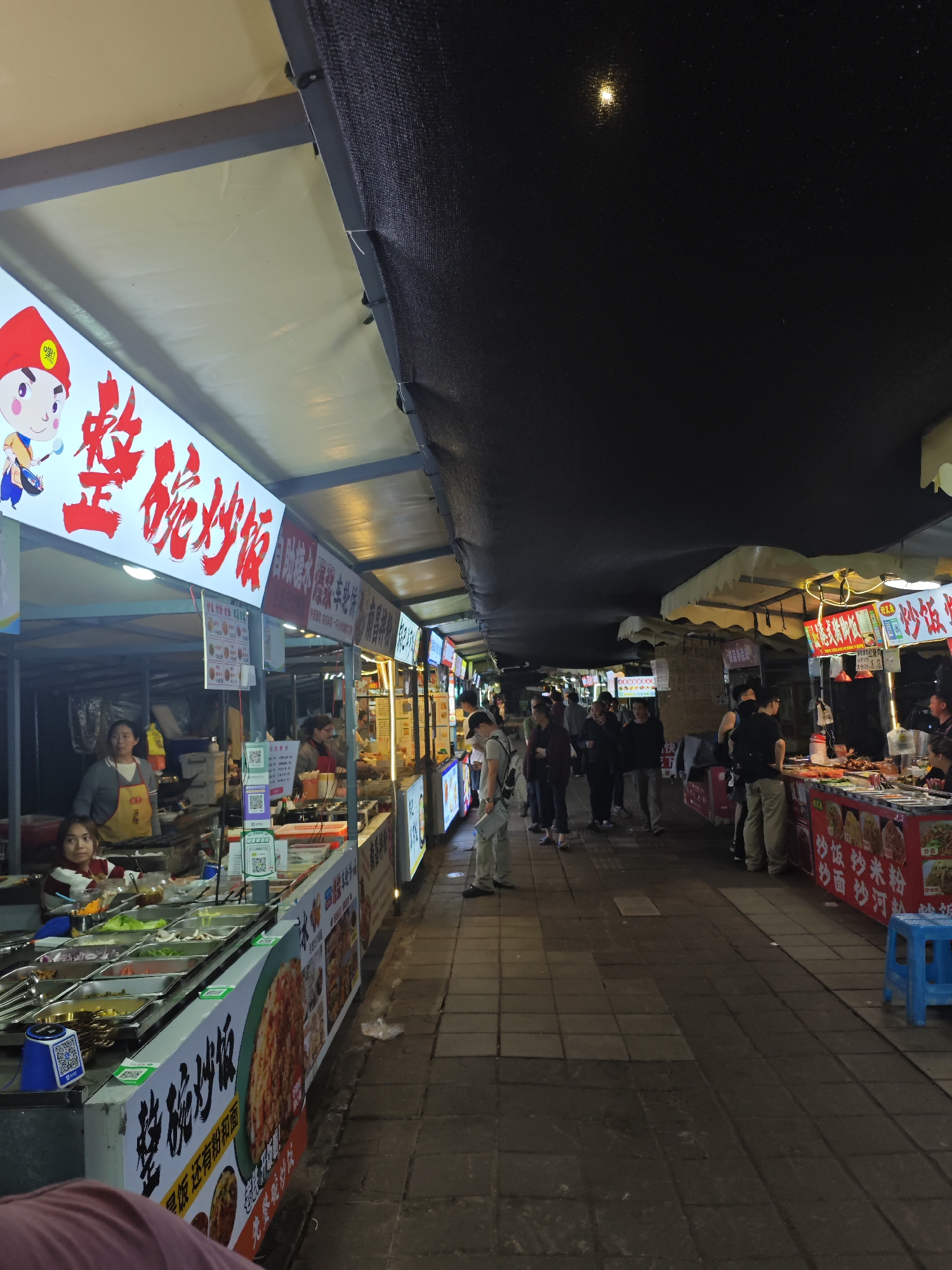 The image captures an outdoor food market, likely in Shapingba District, China. Several stalls line a paved walkway, with black canopies overhead. People are browsing and ordering food. Each stall displays signs with Chinese characters, and images of the dishes served. Food items are visible in containers at the stalls. The lighting suggests it is nighttime.
