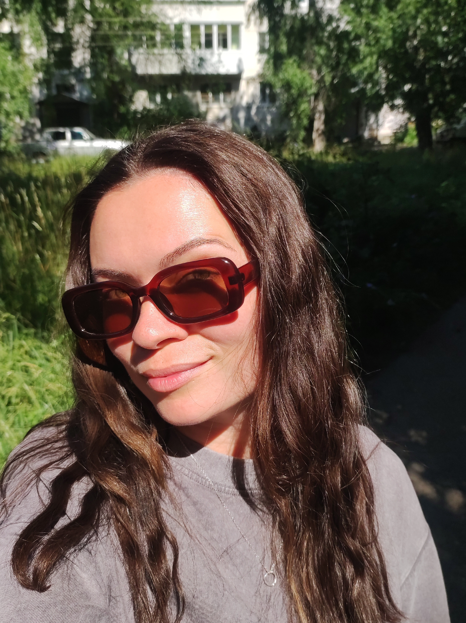 A medium-shot photograph shows a woman with long, dark brown hair wearing large, brown, rectangular sunglasses and a grey crewneck t-shirt. She is standing outdoors in what appears to be a residential area. The background is blurred but shows a building with windows, trees, and green foliage. The sunlight creates a strong shadow across the right side of her face and a glint on her sunglasses. The image quality is good, with natural lighting and moderate detail. The location is not identifiable as Chaikovsky, Russia, or any other specific city.