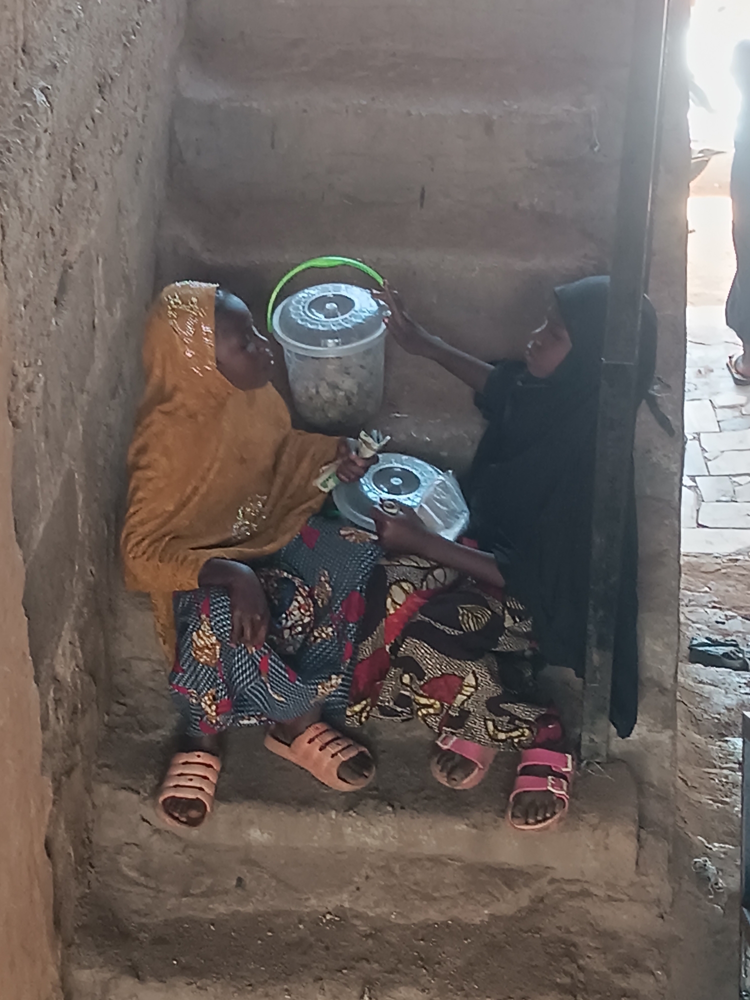 Two young girls are sitting on a set of rough, weathered stone steps. The girl on the left wears a mustard-colored headscarf, a blue patterned dress, and orange sandals. The girl on the right wears a black headscarf and dress, and pink sandals. They are seated close together, interacting with two containers: one is a clear plastic bucket with a green handle, and the other is a smaller, round, clear plastic container. The environment is stark, with walls constructed of rough stone or mud brick. Based on the metadata, the location is Bukuru, Nigeria.
