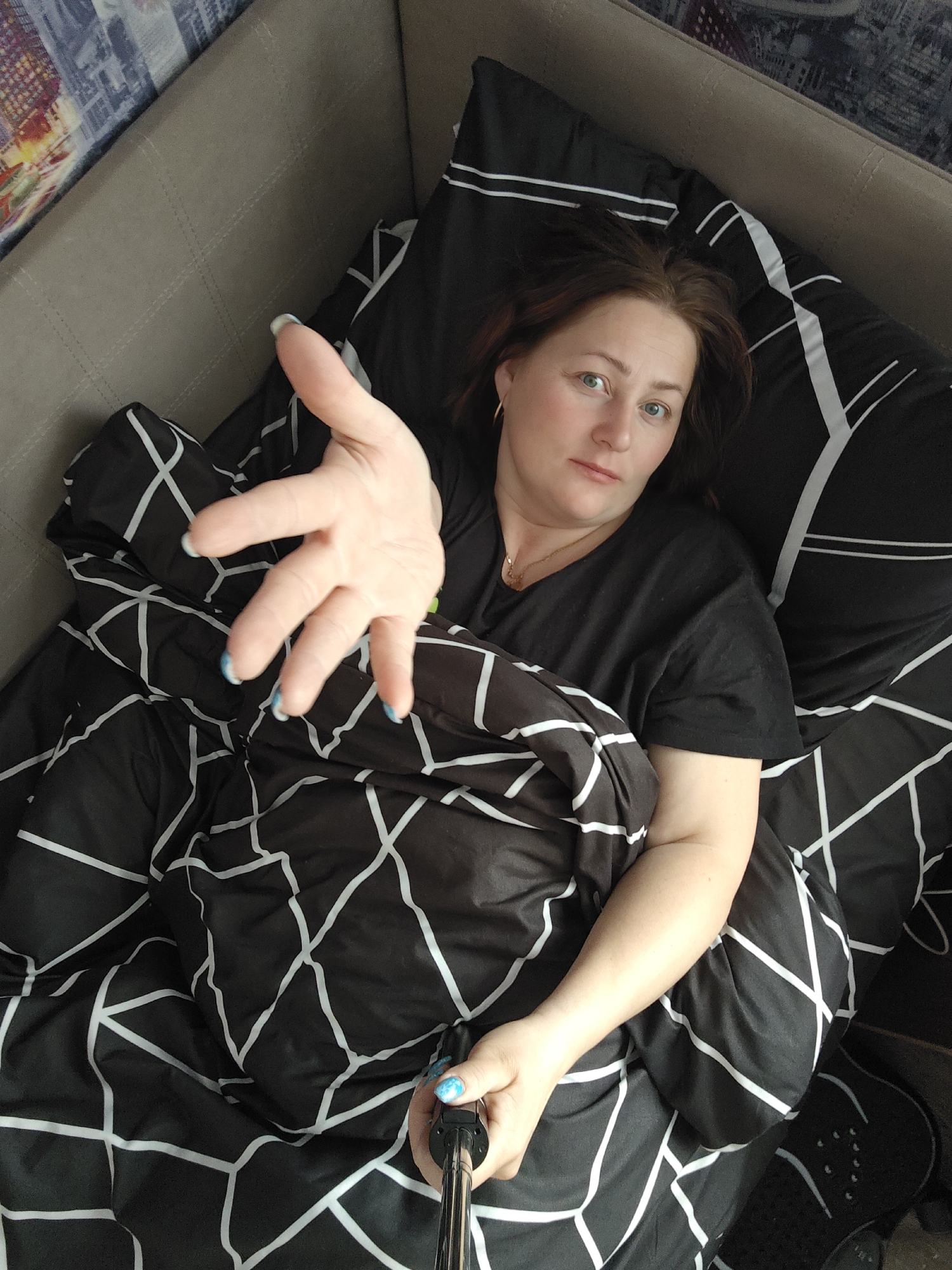 A woman lies in a bed with black and white geometric patterned bedding. She wears a black t-shirt and a gold necklace, and has blue nail polish on her nails. She is holding a selfie stick and reaching out her hand towards the camera. The location is Марковский, Russia.