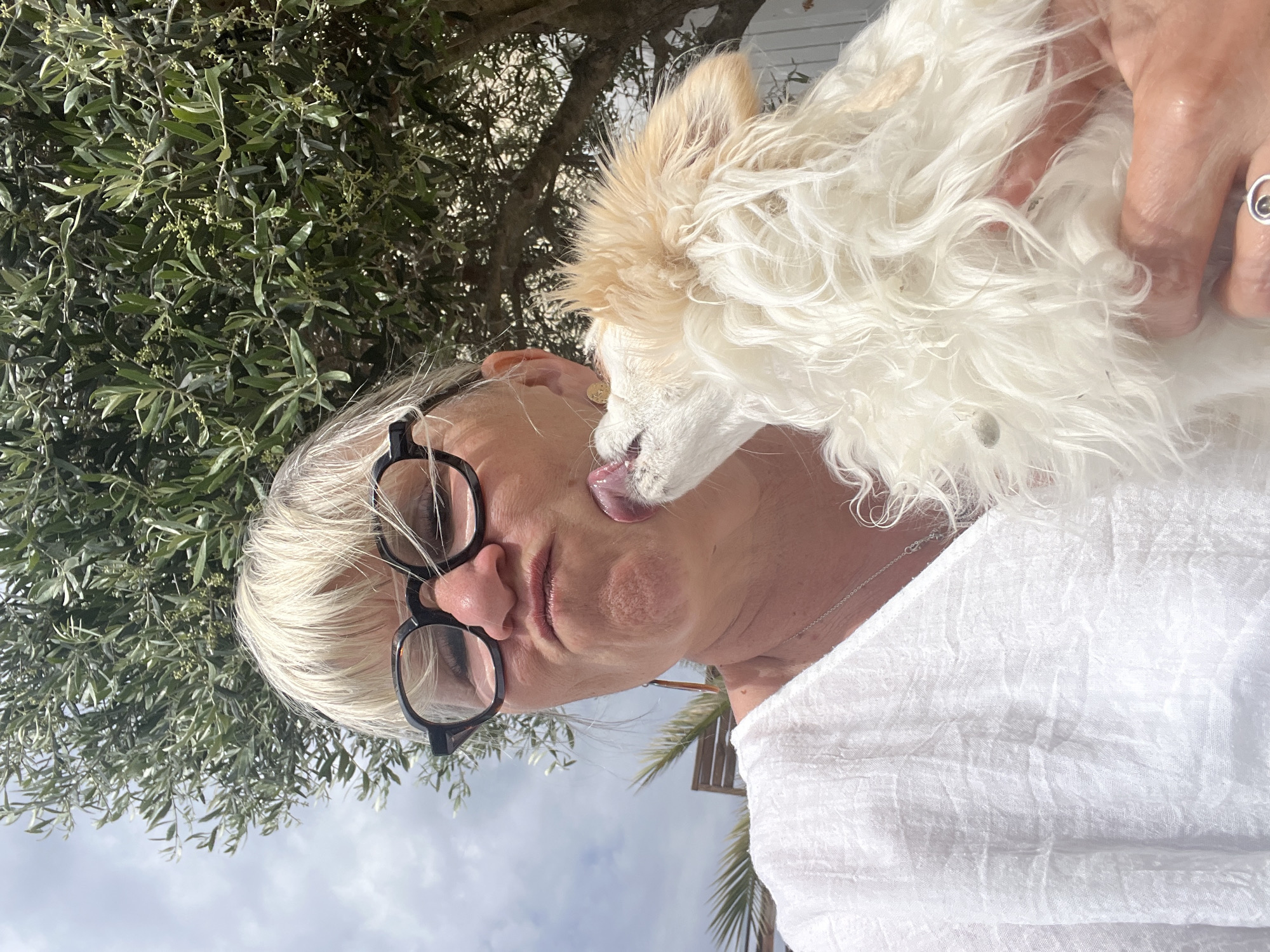 A woman with short, blonde hair, wearing black-rimmed glasses and a white shirt, is holding a small, white and tan dog outdoors. The dog is licking the woman's cheek. An olive tree is visible behind them, with the sky in the background. The photo appears to have been taken in Montarnaud, France, as indicated by the presence of olive trees which are common in that area. A finger with a silver ring on it is also visible.