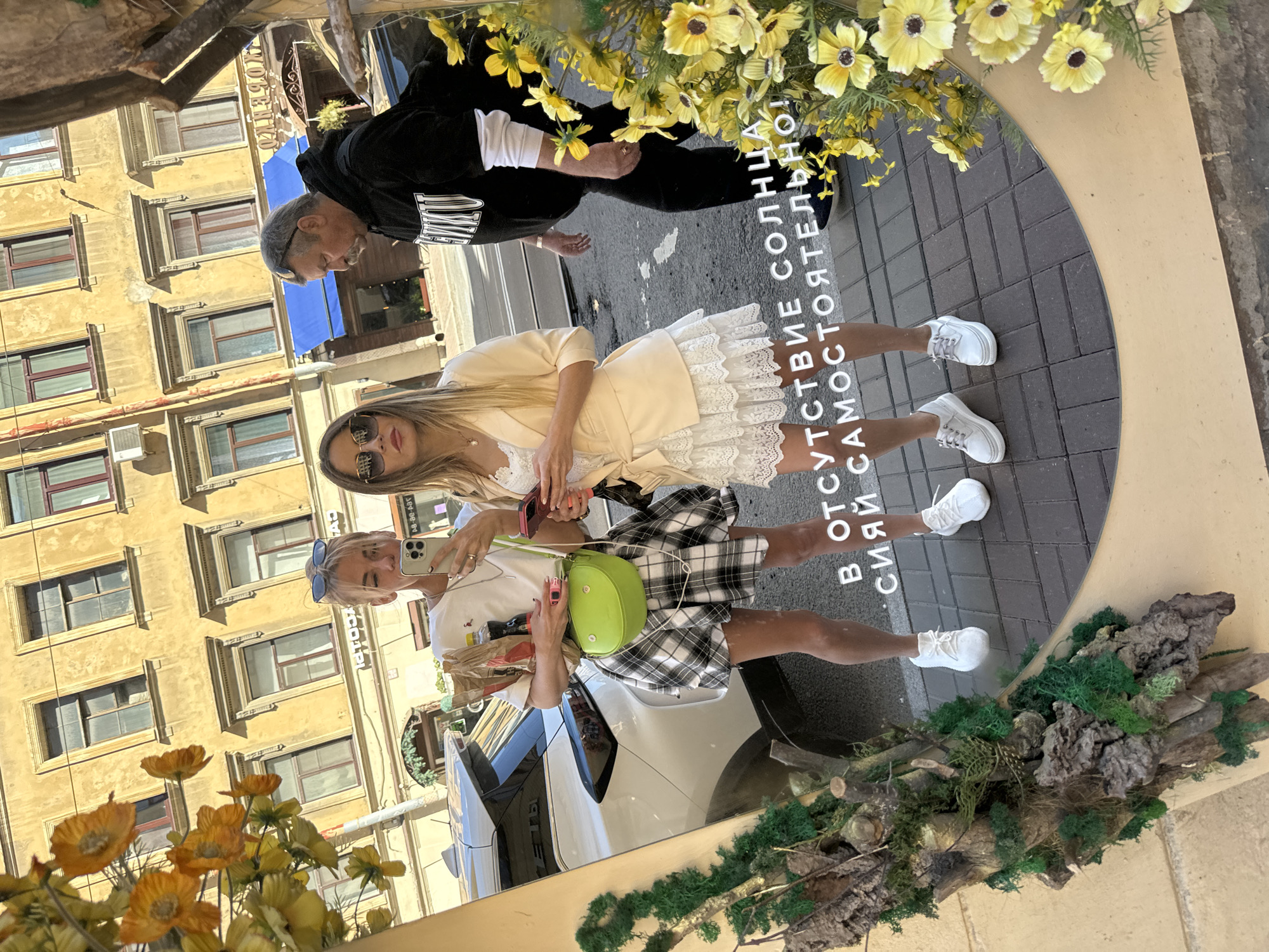 In Saint Petersburg, Russia, two young women are captured in a mirror reflection, taking a selfie on a sunlit street. The woman on the left, with short blonde hair and sunglasses perched on her head, wears a white t-shirt, a black and white plaid skirt, and white sneakers. She holds a phone in her right hand and a shopping bag filled with items in her left. A bright green crossbody bag hangs from her shoulder. The woman on the right, with long blonde hair and sunglasses, sports a white lace dress and a pale yellow blazer, also wearing white sneakers. She holds a phone in her right hand. Behind them, a man in a black hoodie with white lettering walks away from the camera. The background reveals a typical Saint Petersburg street scene with a yellow building adorned with ornate windows and a blue awning. The mirror itself is decorated with artificial greenery and yellow flowers. The text reflected on the pavement reads, "В ОТСУТСТВИЕ СОЛНЦА СИЯЙ САМОСТОЯТЕЛЬНО!" which translates to "In the absence of sun, shine on your own!"